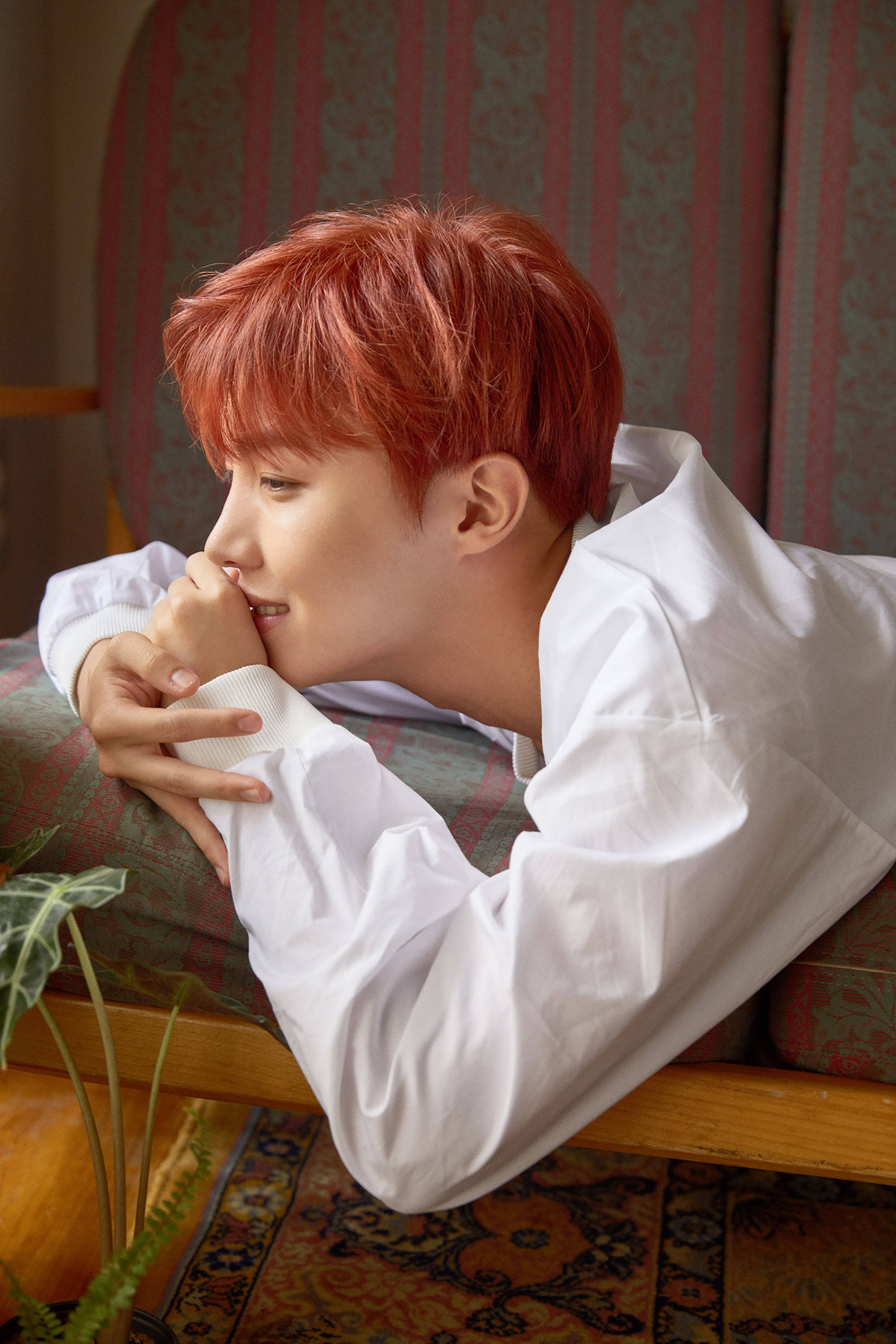 J-hope In Red Hair 壁紙 — 一位年輕男子擁有鮮豔的紅髮，在溫暖照明的房間裡，將下巴擱在交疊的雙手上，懶洋洋地躺在圖案沙發上。