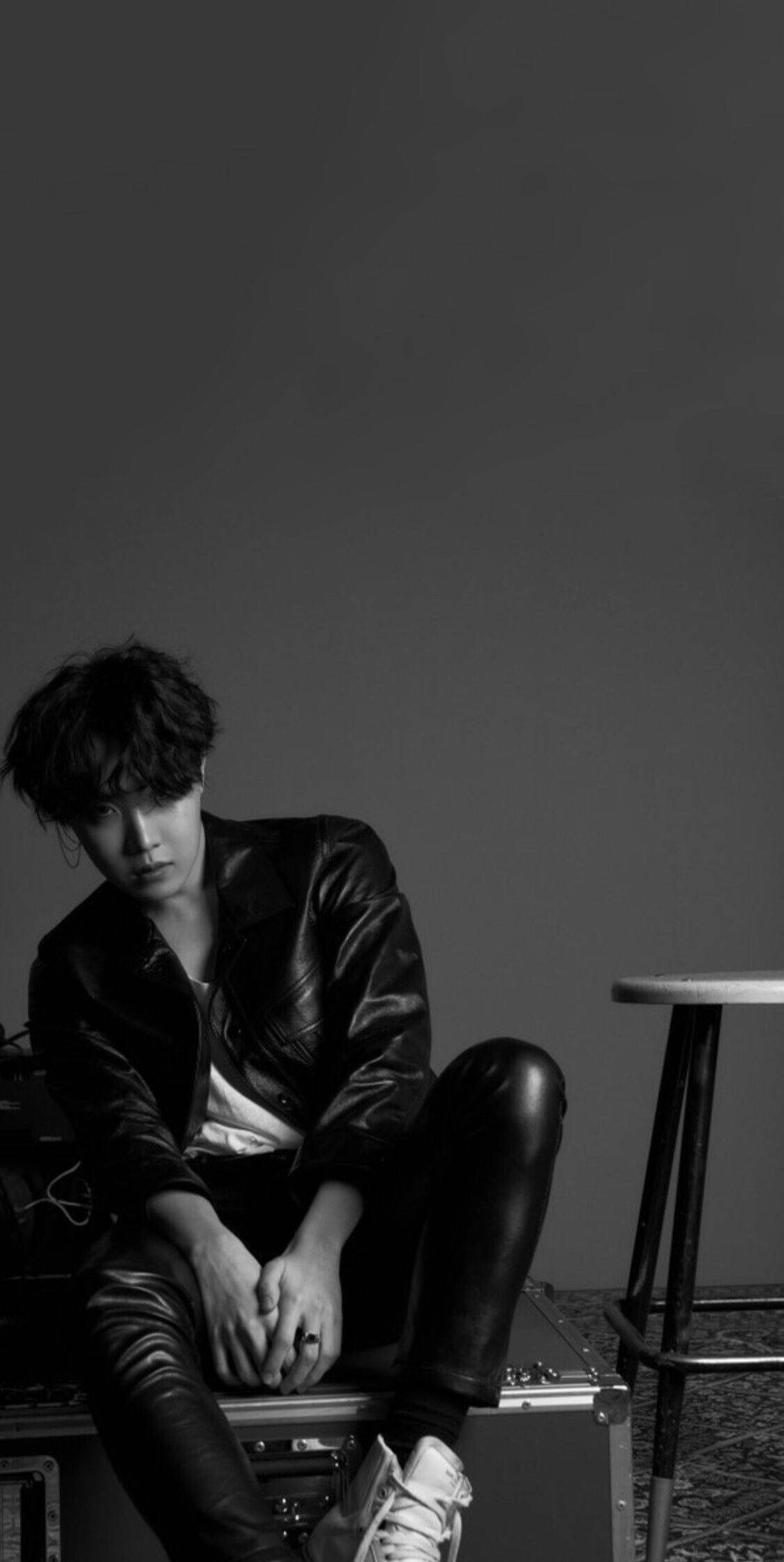 J-hope Love Yourself Tear 壁紙 — 一位身穿皮褸的年輕男子，在單色工作室拍攝中，沉思地坐在一個齒輪箱上，背景是純灰色的牆壁。
