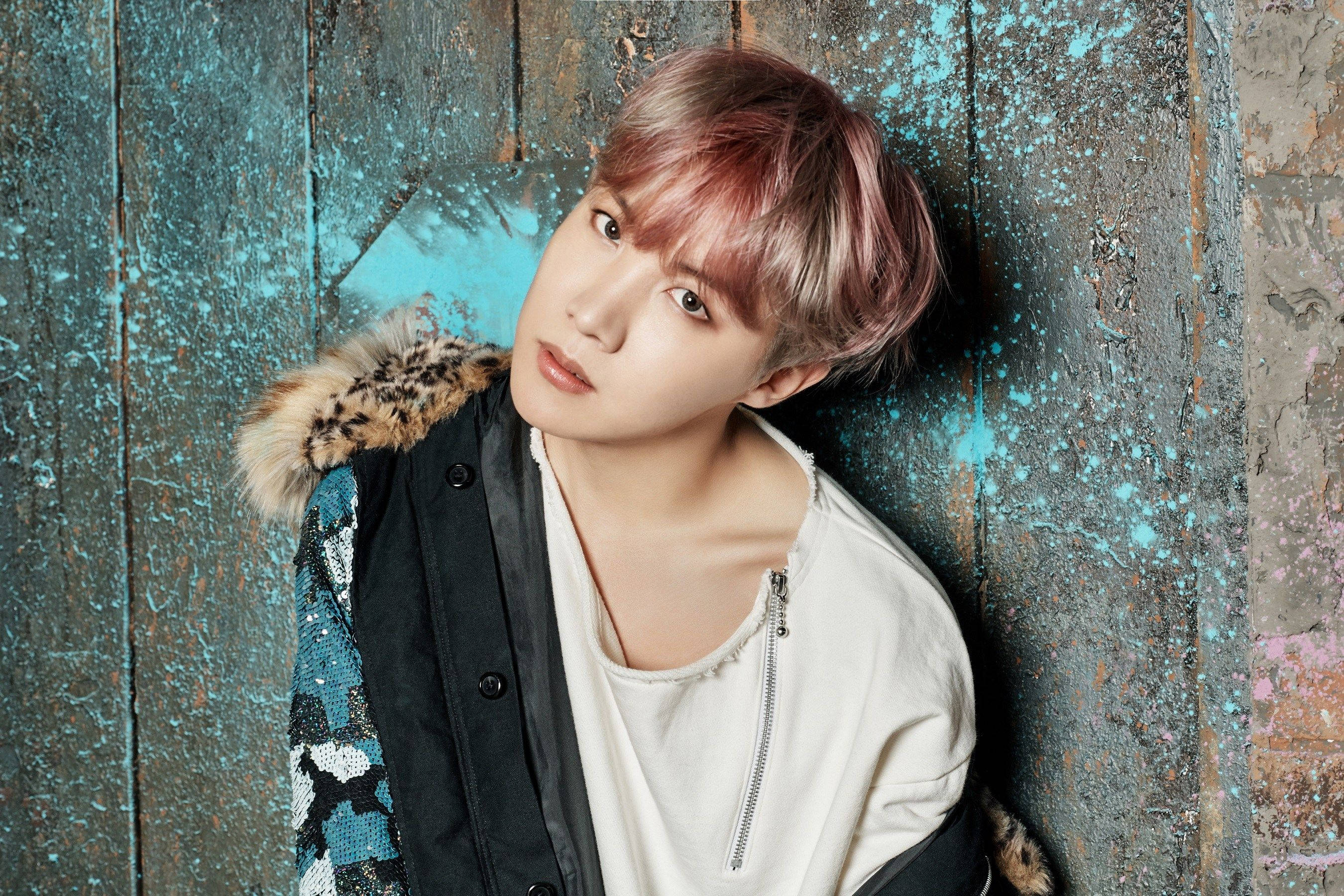 J-Hope 可愛概念照 壁紙 — 一位粉紅色頭髮的年輕人靠著風化的藍色木牆，身穿帶有豹紋毛邊的夾克。