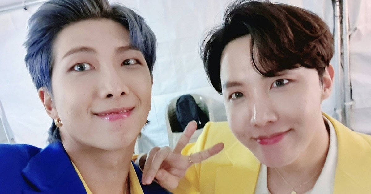 J-Hope 與 Rap Monster 合照 壁紙 — 兩位微笑的男士，一頭藍色和棕色頭髮，在明亮的室內環境中緊密擺姿勢，其中一人做出和平手勢。