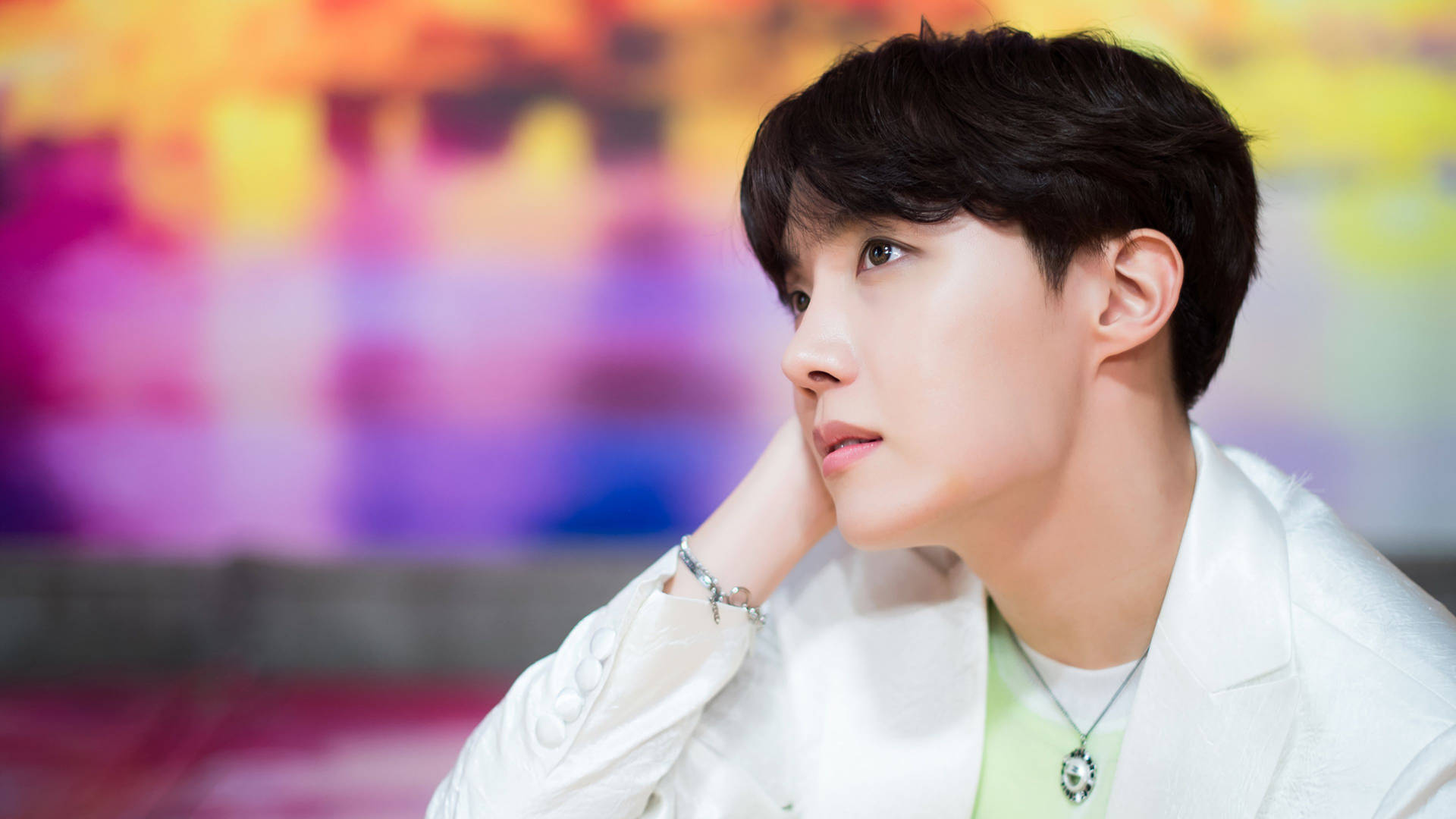 J-Hope夢幻風 壁紙 — 一位身穿白色外套的年輕男子，在充滿活力的模糊彩虹背景下若有所思地凝視。