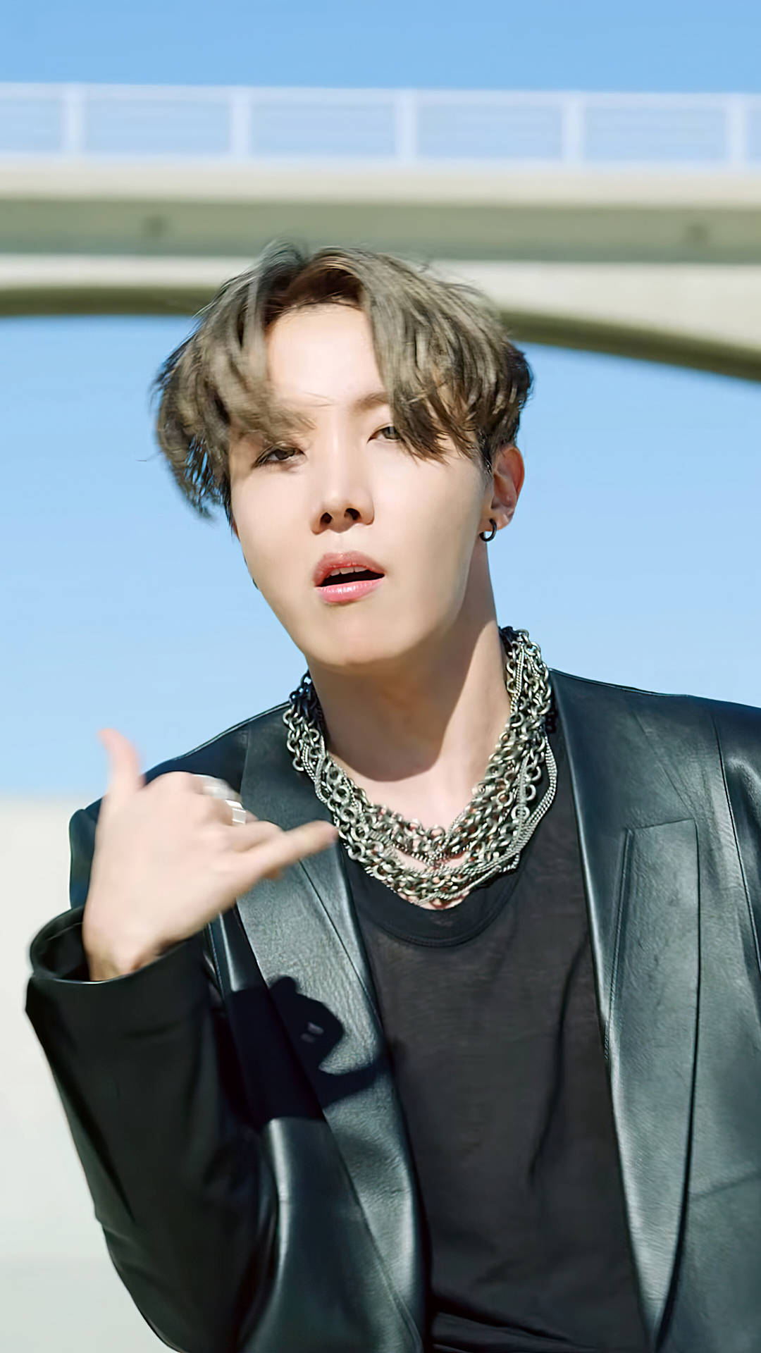 J-Hope帥氣亮相 壁紙 — 一位年輕男子，梳著時尚髮型，佩戴多層銀鏈，在橋下以清澈藍天為背景自信地擺姿勢。