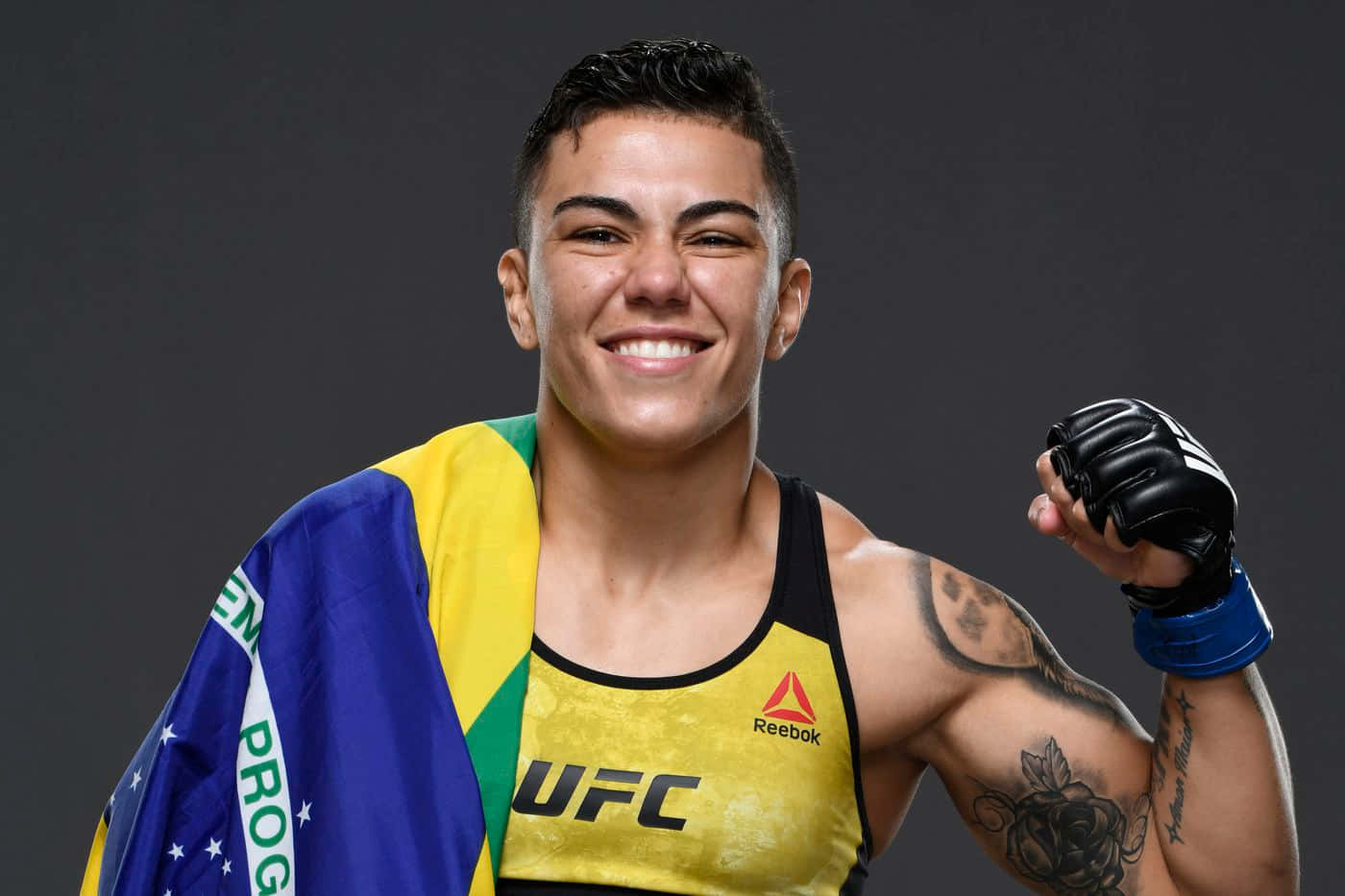 巴西女性MMA拳手微笑著舉起戴手套的拳頭，肩上披著國旗。