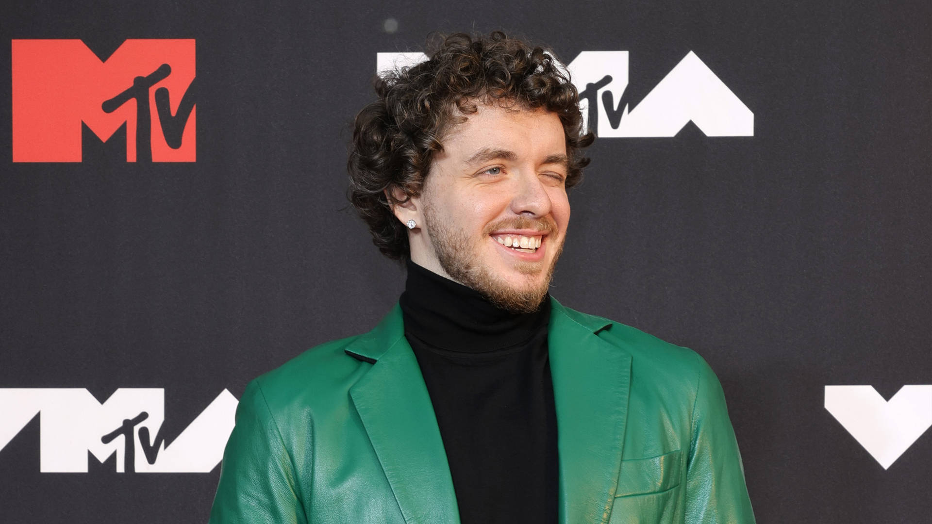 Jack Harlow 在 MTV 頒獎典禮 壁紙 — 一位笑容滿面、捲髮的男子在MTV活動中穿著綠色皮褸和黑色高領毛衣。