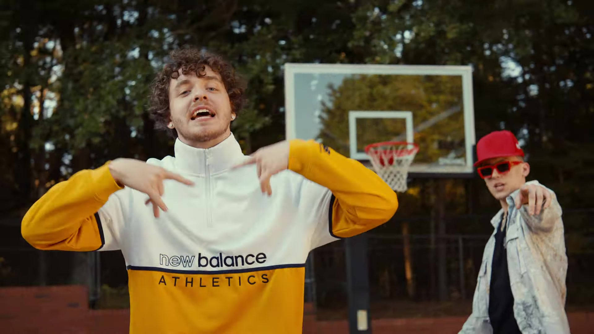 Jack Harlow Tyler Herro MV 照片 壁紙 — 兩名年輕男子在戶外籃球場上充滿活力地擺姿勢，背景有樹木和籃框。