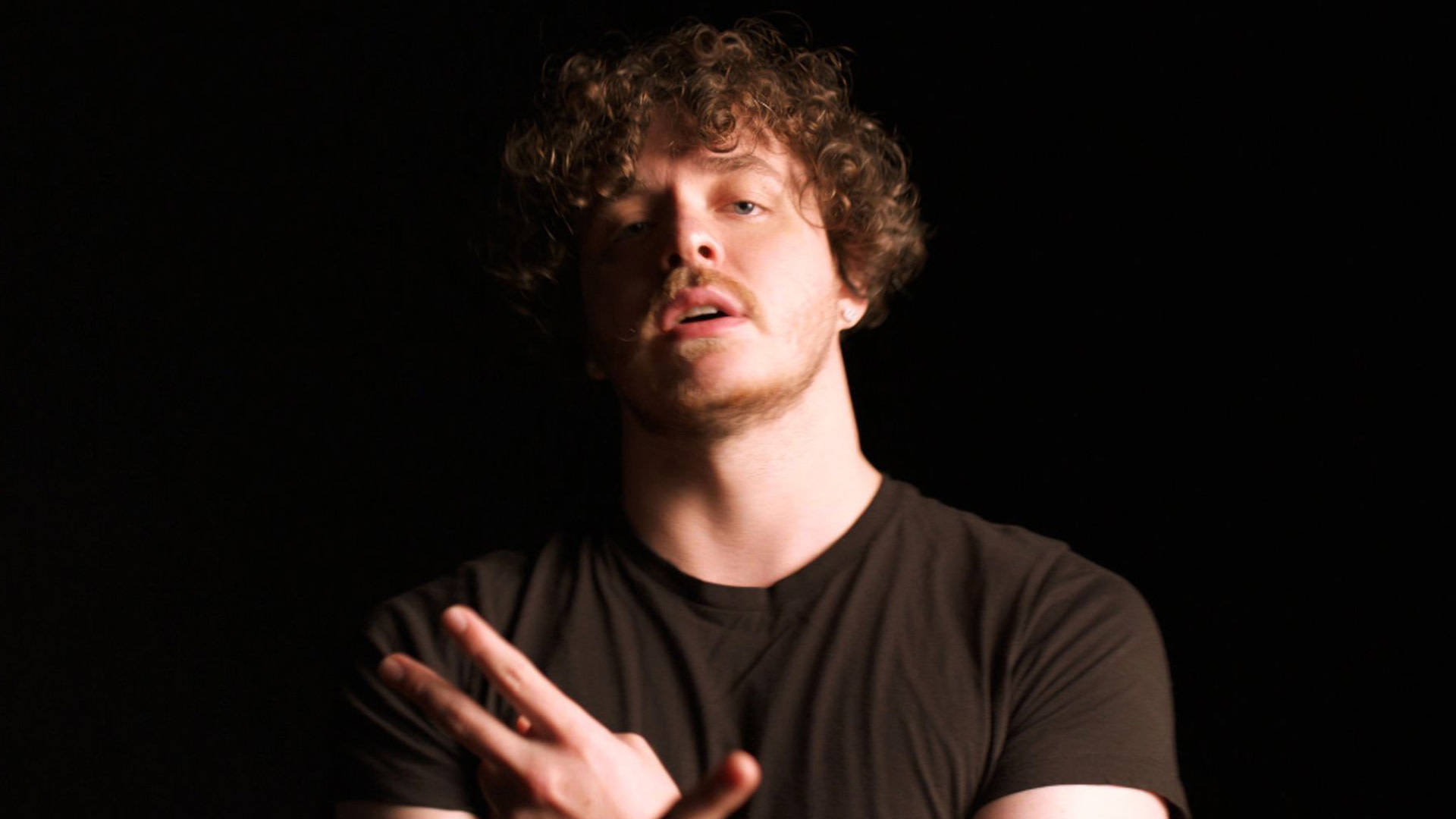 Jack Harlow 黑幕布前 壁紙 — 一個留著捲髮和鬍鬚的男人在戲劇性燈光下對著純黑色背景做手勢。