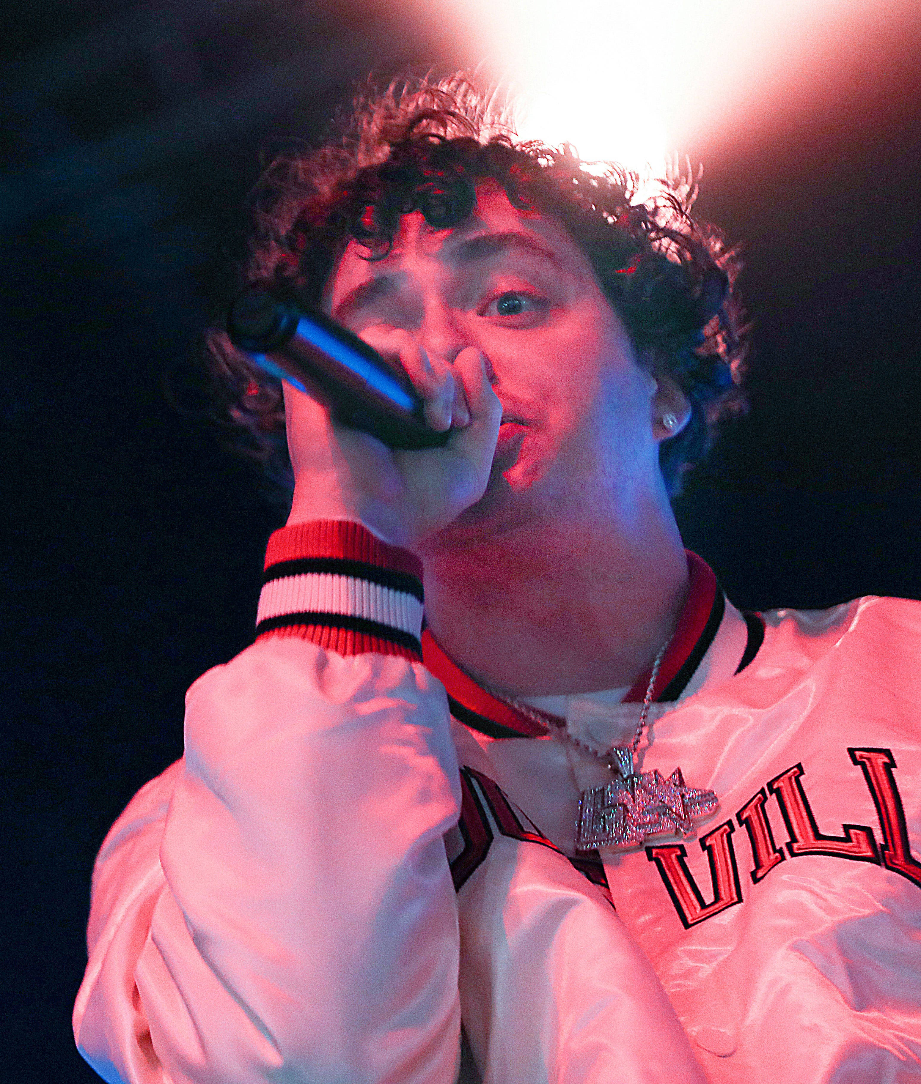 Jack Harlow 手持麥克風 壁紙 — 一位捲髮年輕男子在戲劇性的紅藍舞台燈光下，穿著閃亮白色外套對著麥克風唱歌。