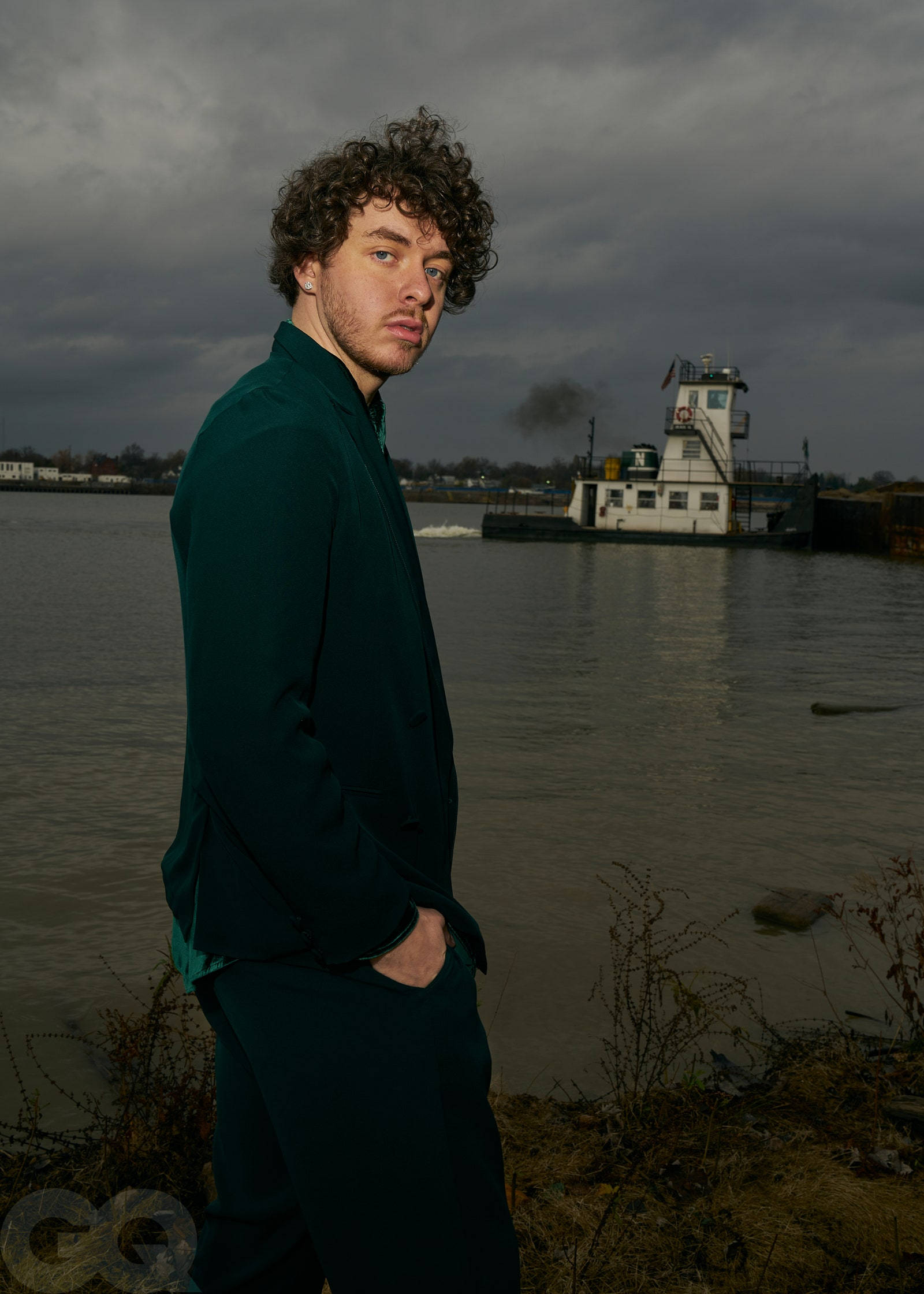 Jack Harlow 湖畔 壁紙 — 一名身穿深綠色服裝的男子站在河岸邊，天空多雲，背景有一艘拖船。