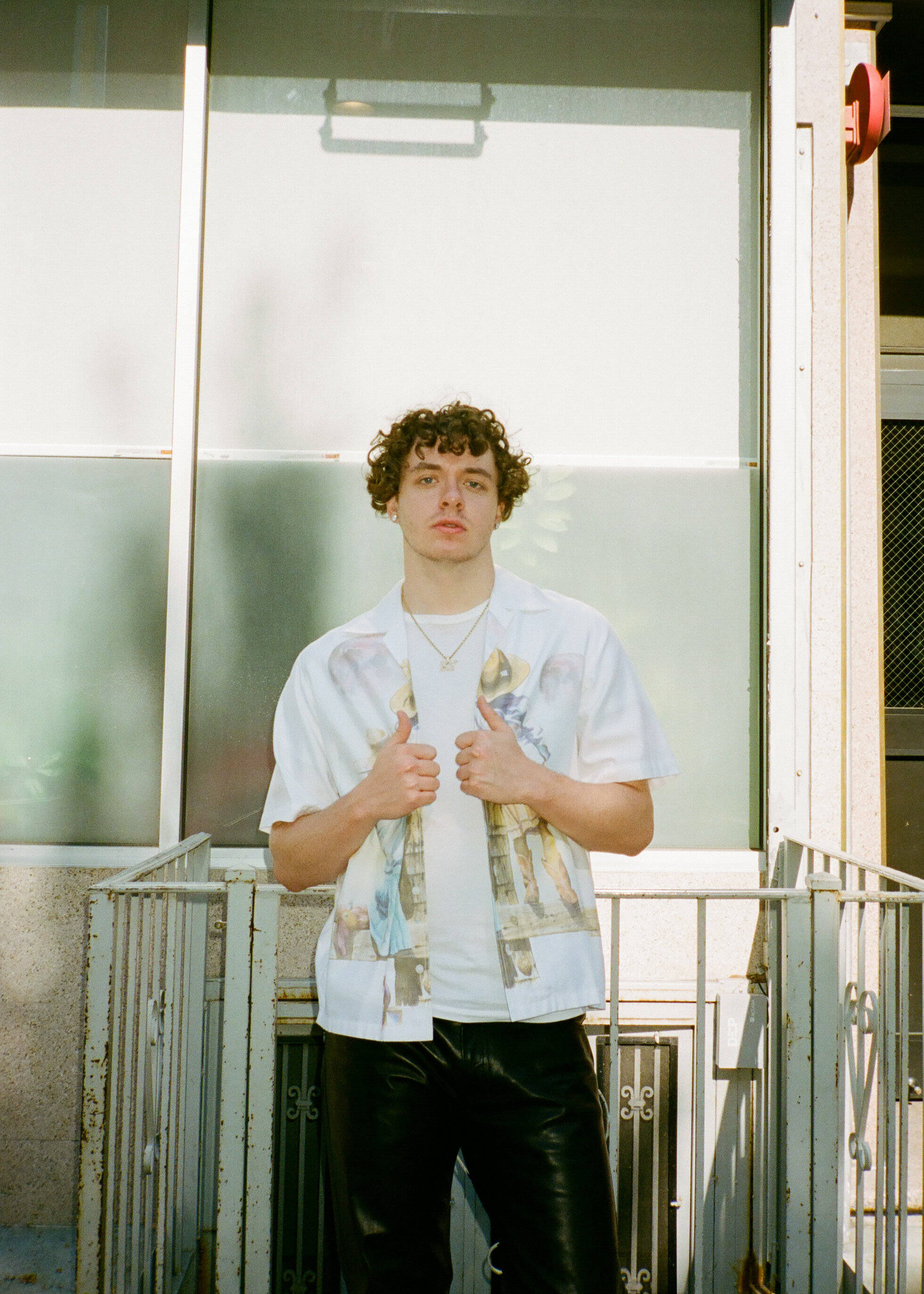 Jack Harlow 肖像照 壁紙 — 一位捲髮年輕男子站在陽光普照的陽台上，穿著圖案T恤和黑色長褲，豎起兩個大拇指。