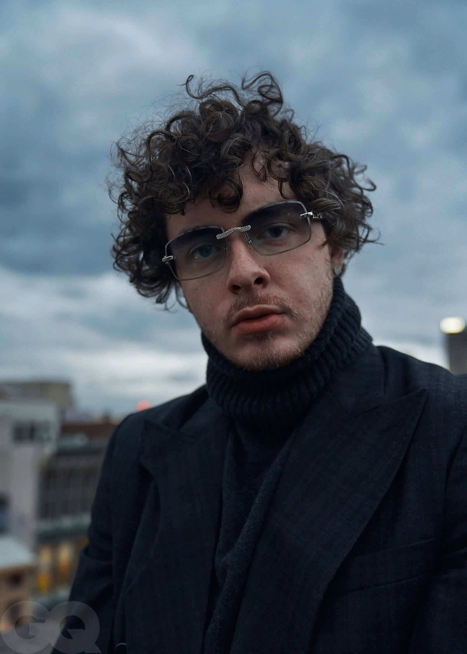 Jack Harlow 自拍 壁紙 — 一個捲髮戴眼鏡的男人在黃昏時分凝視著多雲的城市天際線。