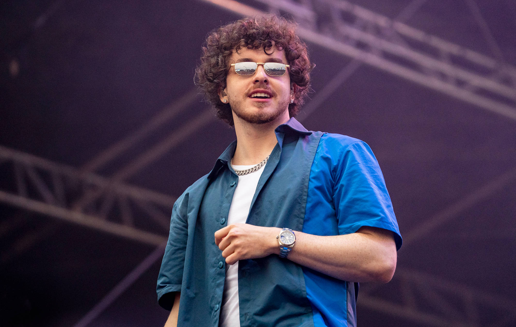 Jack Harlow 舞台演出照 壁紙 — 一個有曲髮和太陽眼鏡的男人，在紫色燈光下穿著藍色襯衫和鏈條在舞台上表演。