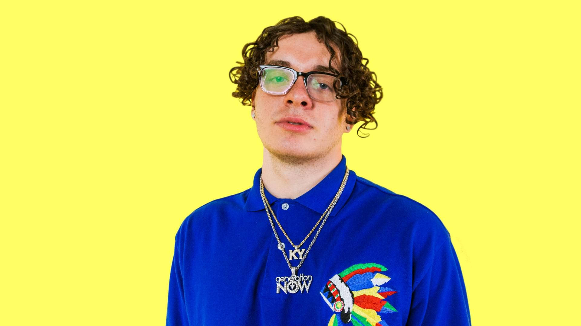 Jack Harlow 黃色主題照 壁紙 — 一位捲髮戴眼鏡的年輕男子，身穿帶有彩色刺繡的藍色Polo衫，背景為明亮的黃色。