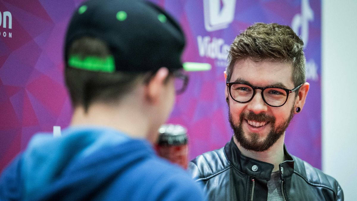 Jacksepticeye VidCon 活動照 壁紙 — 一位戴眼鏡、留鬍鬚的男士在充滿活力的紫色品牌活動背景前與另一人交談。