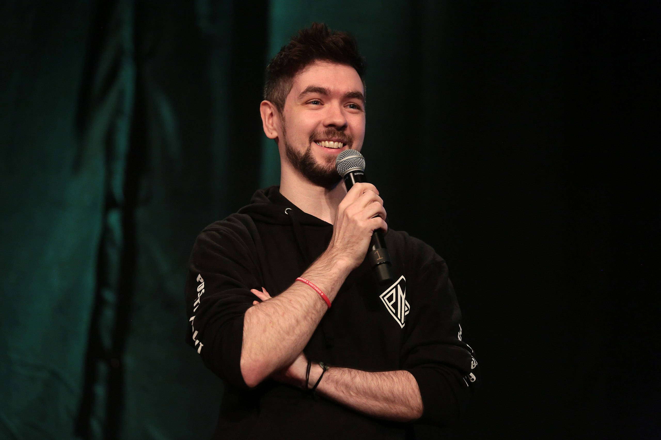 Jacksepticeye 演出現場 壁紙 — 一位留鬍鬚的男士面帶微笑，在深綠色窗簾背景下於舞台上手持麥克風。