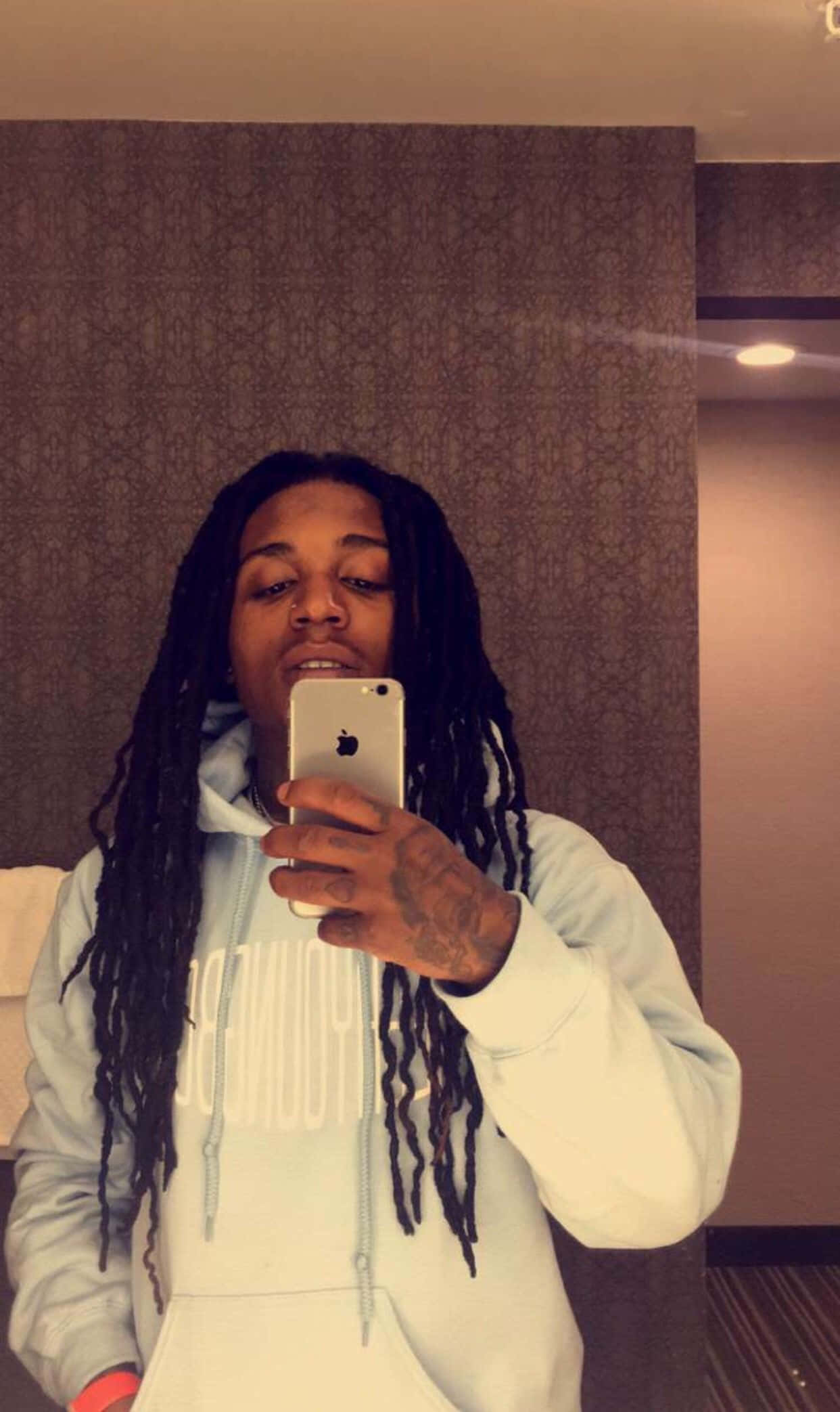 Jacquees 鏡前自拍 壁紙 — 一位留著長髮辮的年輕男子在酒店房間穿著淺藍色連帽衫對著鏡子自拍。
