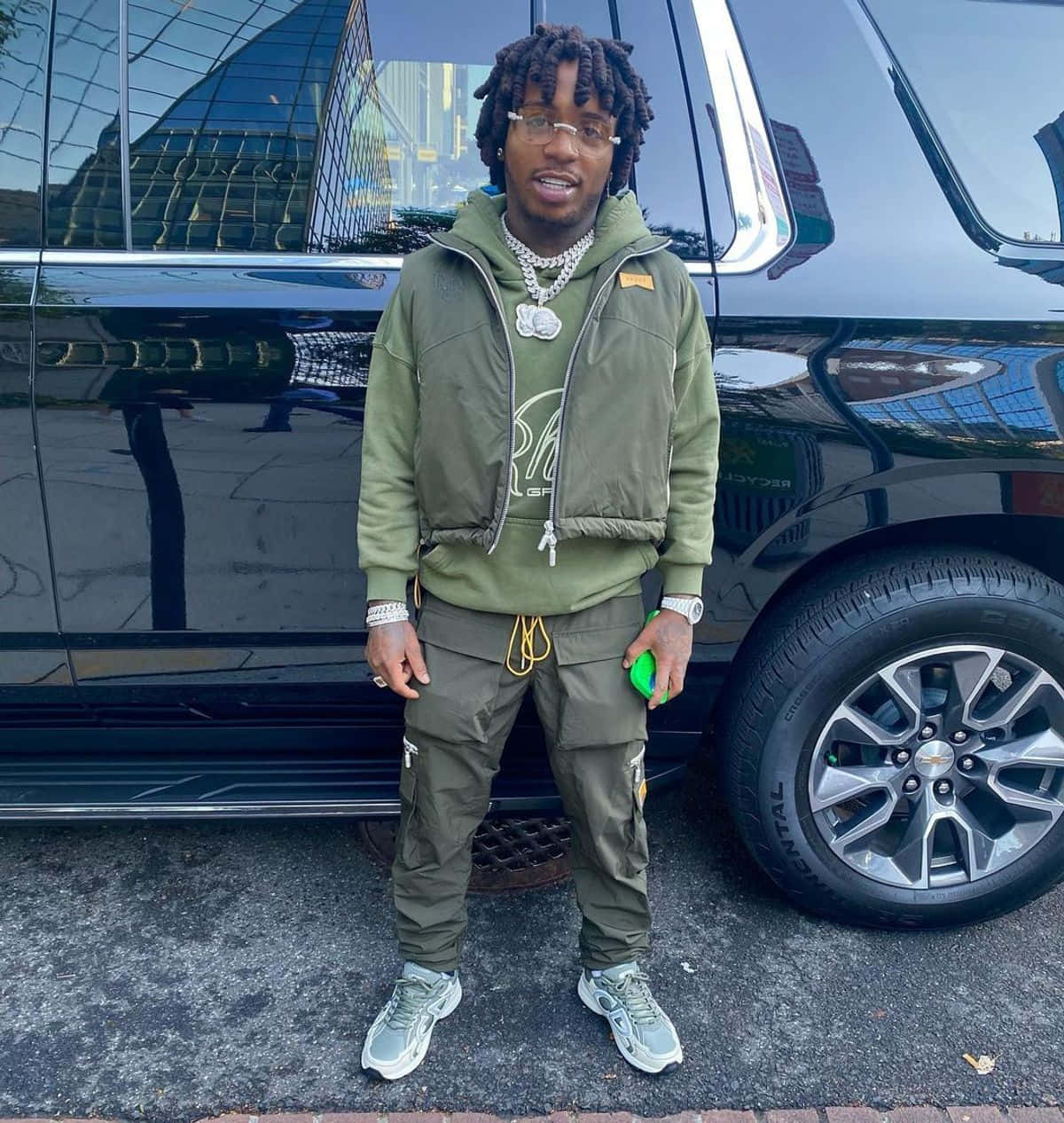 Jacquees 軍綠色造型 壁紙 — 一位時尚男士留著辮子頭、戴著眼鏡，自信地站在城市環境中一輛亮黑色SUV旁。
