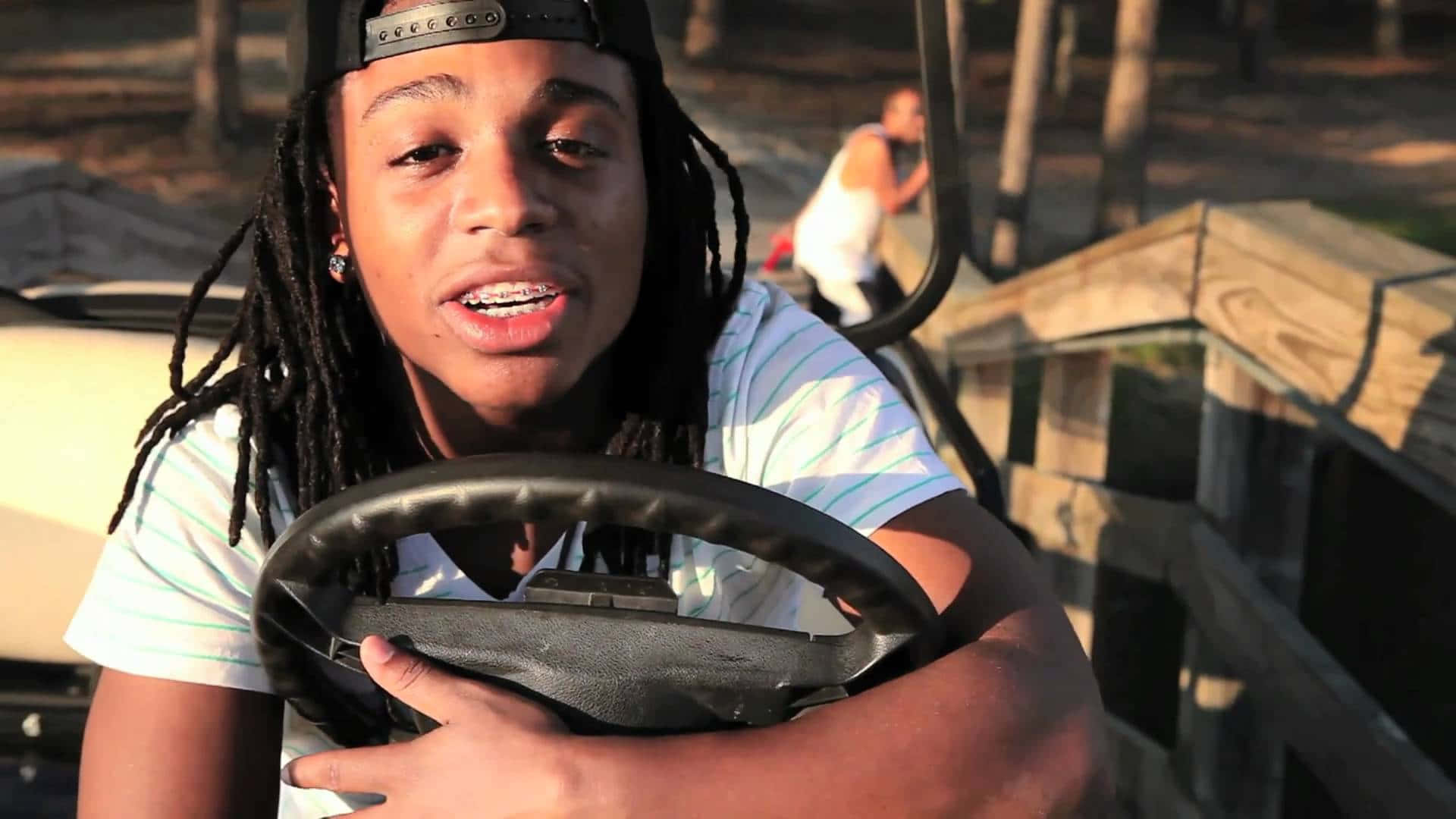 Jacquees 舞台聚光燈下 壁紙 — 一位留著辮子的年輕人在車內握著方向盤，陽光穿過他們身後的樹木照射進來。