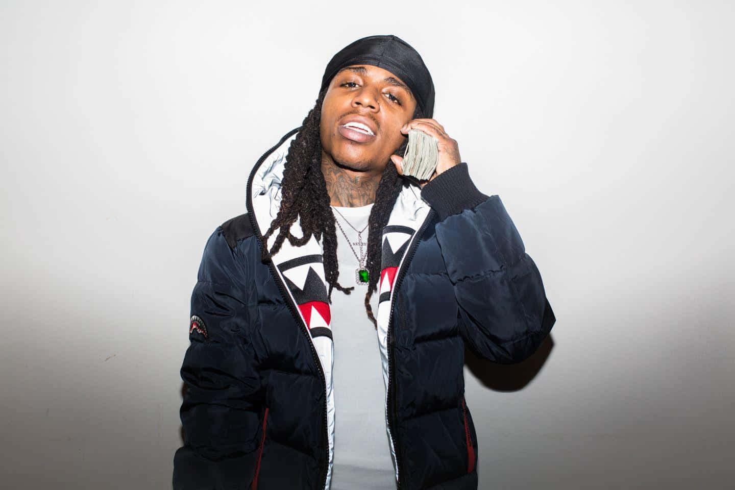 Jacquees 風景攝影寫真 壁紙 — 一名留著雷鬼辮、戴黑色冷帽的男子，在純白背景前將一疊現金像電話一樣貼在耳邊。