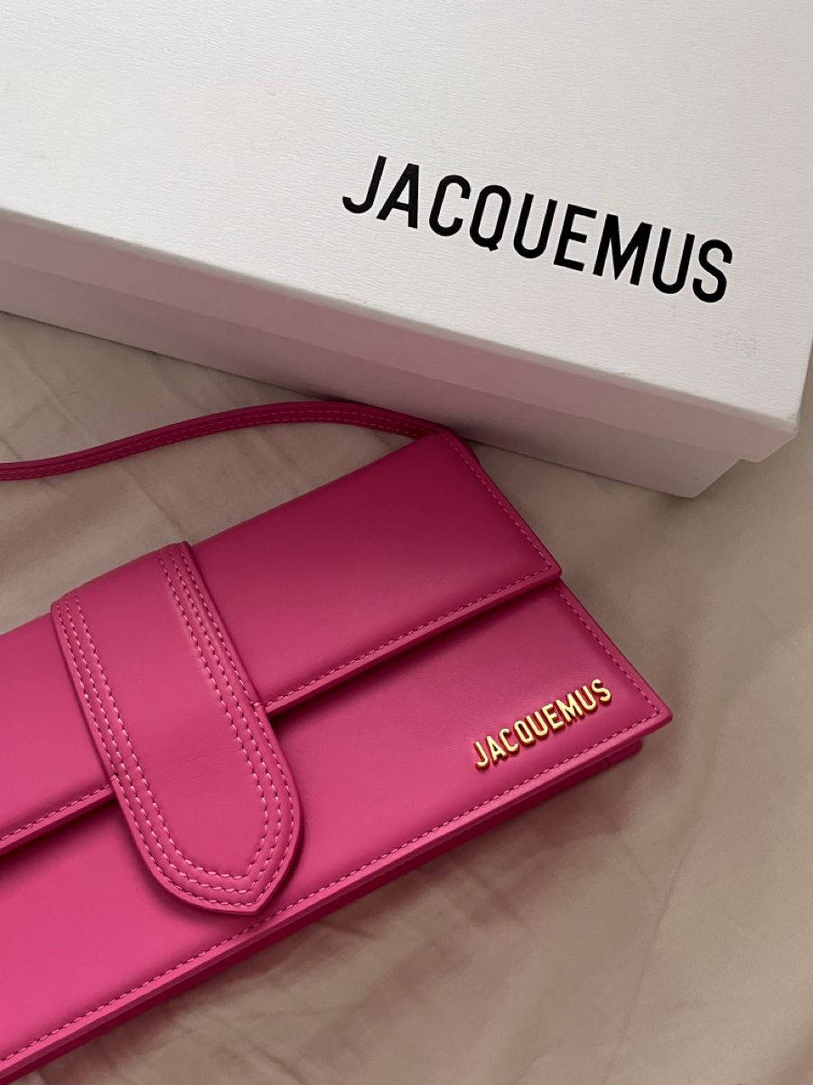 Jacquemus 粉紅 Le Bambino 手袋 壁紙 — 一個充滿活力的粉紅色Jacquemus錢包放在其品牌白色盒子旁邊，置於柔軟的米色表面上。