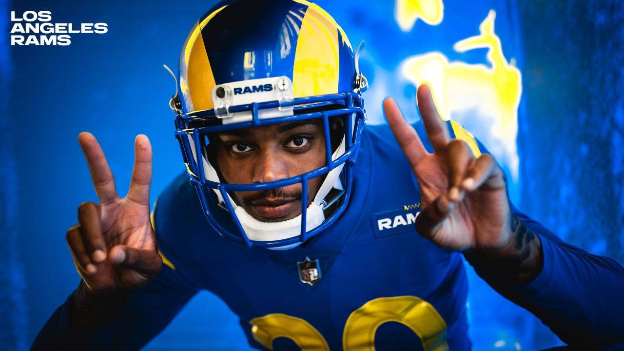 Jalen Ramsey 和平姿勢 壁紙 — 一位身穿藍色和金色裝備的洛杉磯公羊隊美式足球員在發光的藍色背景前做出和平手勢。