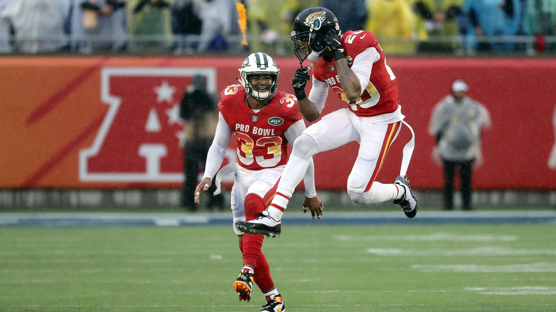 Jalen Ramsey 與 Jamal Adams 壁紙 — 兩名身穿紅白球衣的美式足球員在雨中職業碗比賽中，在綠色場地上動態跳躍。