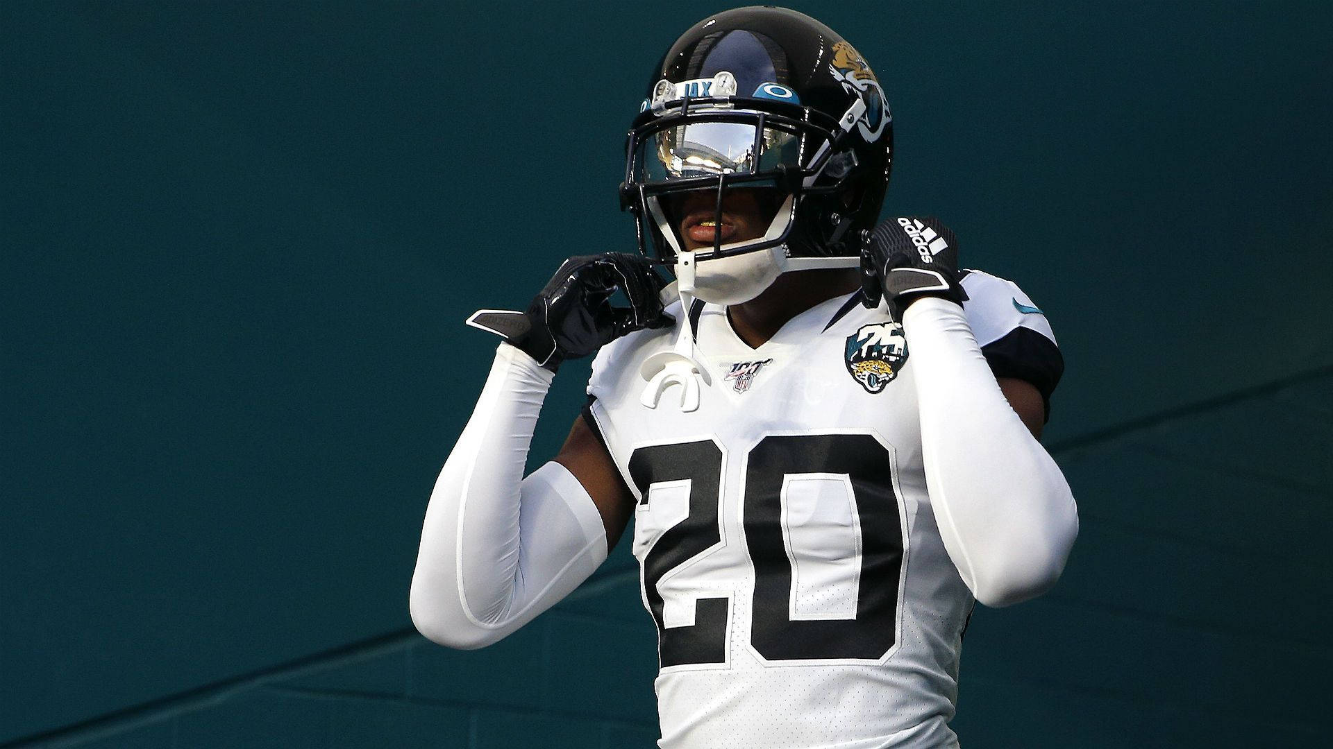 Jalen Ramsey 賽場全神貫注 壁紙 — 一名傑克遜維爾美洲虎隊的美式足球員，身穿白色20號球衣，在青綠色牆壁旁調整頭盔。