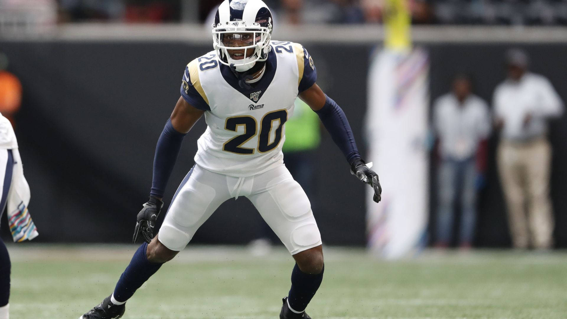 Jalen Ramsey 賽場動態 壁紙 — 一名身穿白色和藍色公羊隊球衣、號碼20的美式足球員在比賽期間警覺地站在綠色場地上。