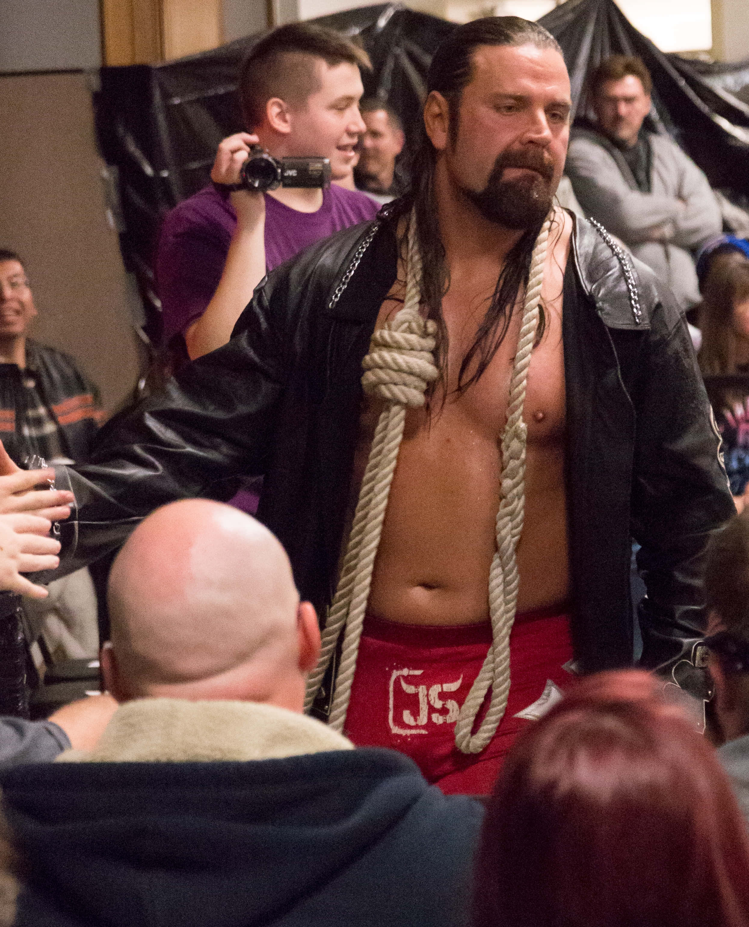 James Storm於Empire State Wrestling賽事中激烈比賽 壁紙 — 一名肌肉發達的摔角手，脖子上纏著繩子，身穿黑色皮褸和紅色短褲，站在舞台中央，被觀眾包圍。