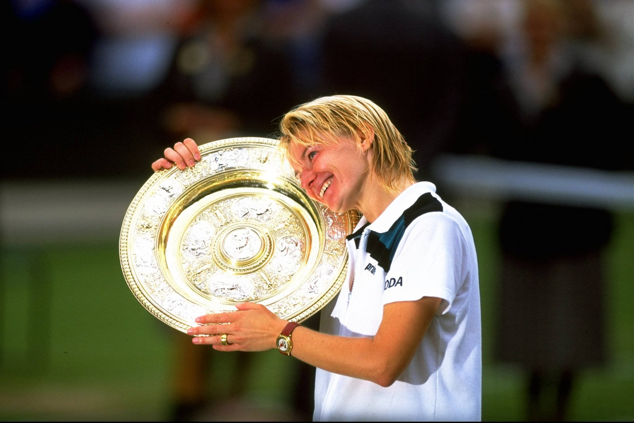 Jana Novotna 手捧獎盃特寫 壁紙 — 一位快樂的金髮網球選手在綠色球場上手持一個華麗的大銀獎盃，笑容燦爛。