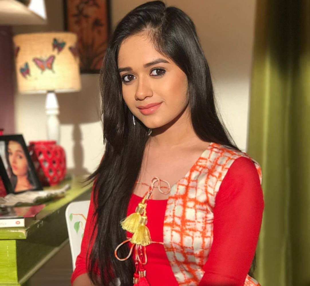 Jannat Zubair 身著傳統印度服飾展現優雅 壁紙 — 一位長黑髮的年輕女性在室內靠近蝴蝶燈和綠色窗簾的地方坐著，溫柔地微笑。