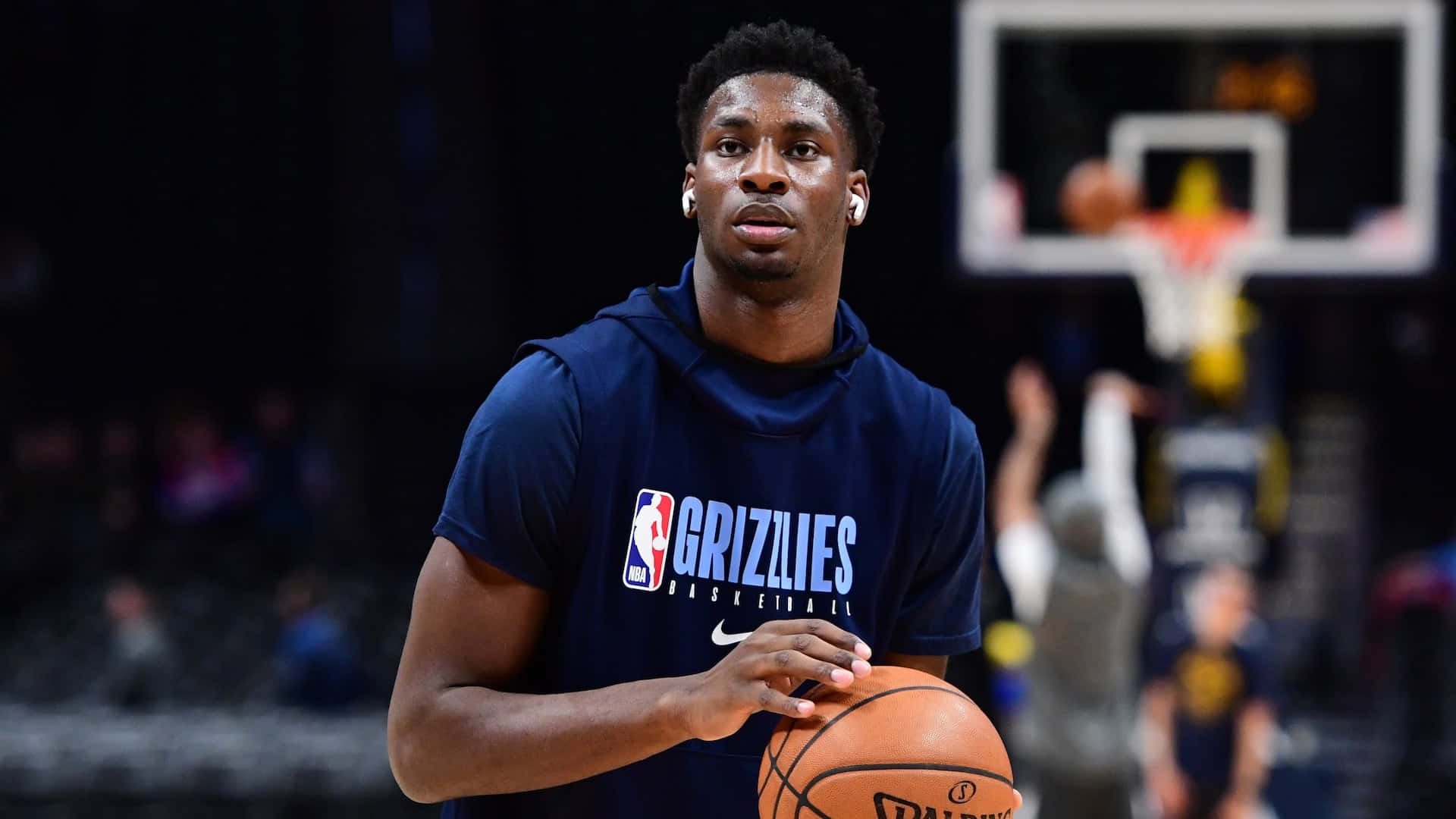 Jaren Jackson Jr. NBA 訓練 壁紙 — 一位專注的籃球運動員身穿孟菲斯灰熊連帽衫，在場館燈光下持球，身後是模糊的球場。