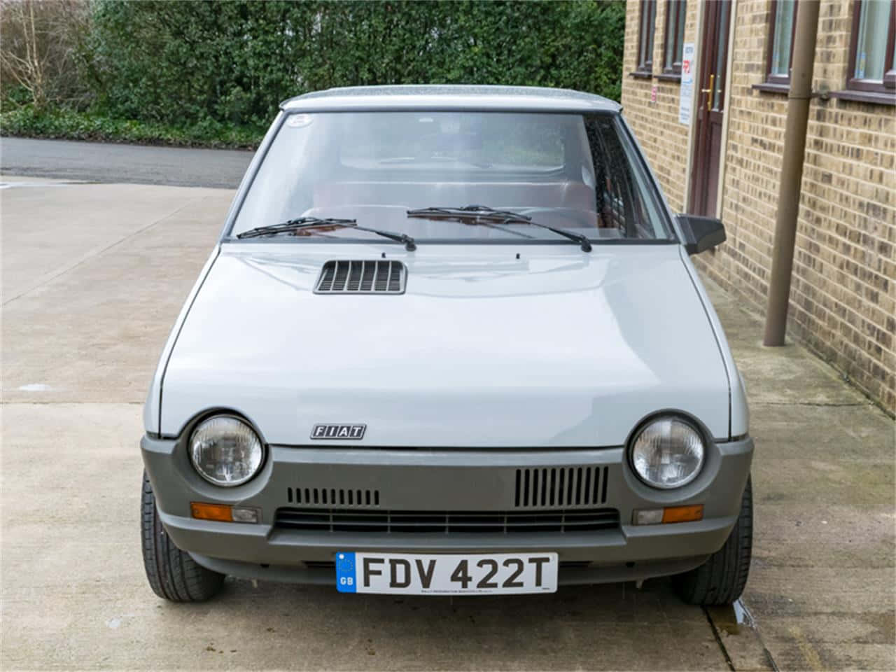 Fiat Ritmo 桌布 壁紙 — 一架銀色嘅Fiat 126泊喺磚樓旁邊嘅混凝土車道上，背景有綠色灌木。