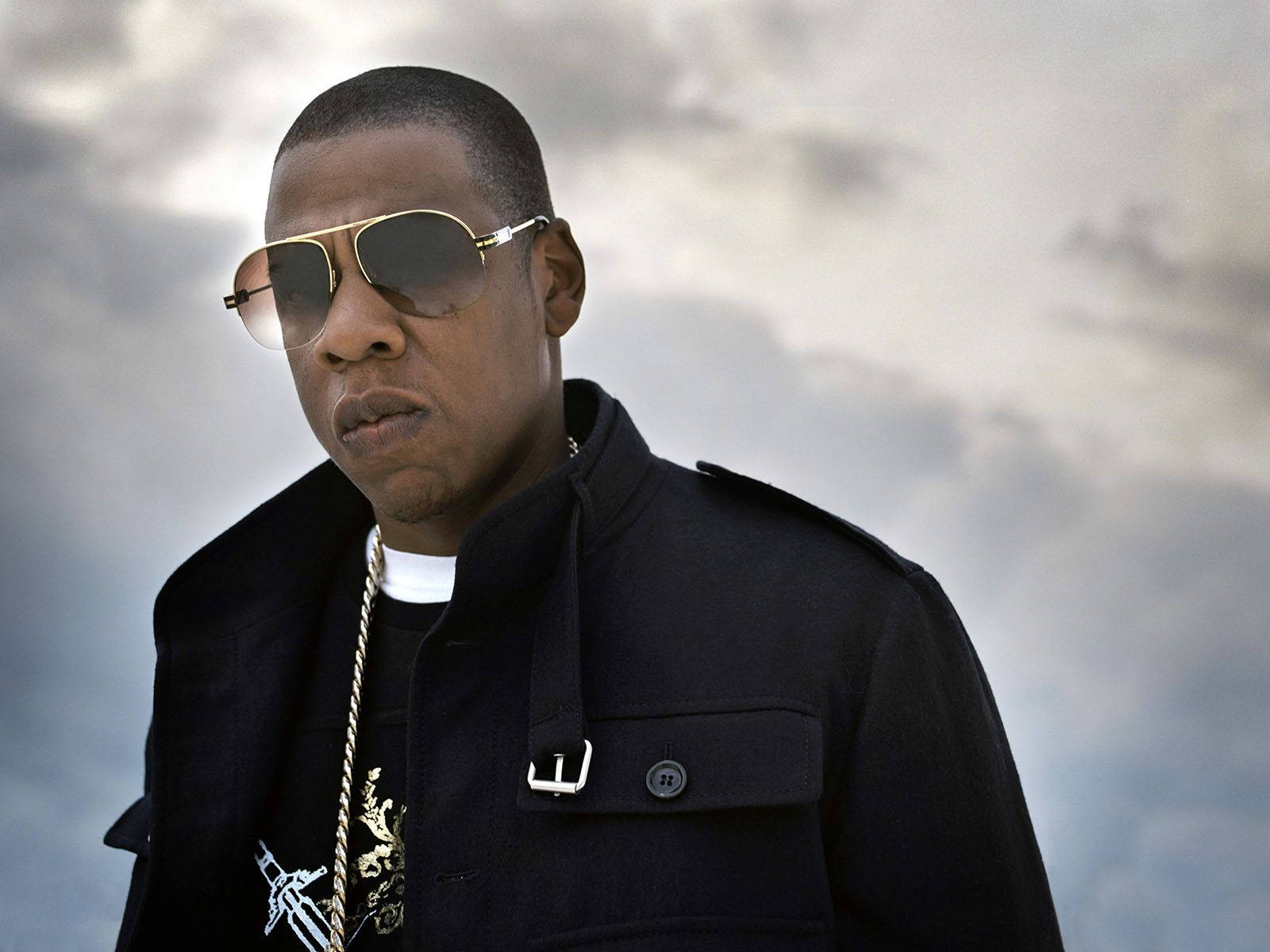 Jay-Z 真實肖像 壁紙 — 一個穿著黑色外套和金色邊框太陽眼鏡的男人，在陰天的天空下帶著嚴肅的表情站立。