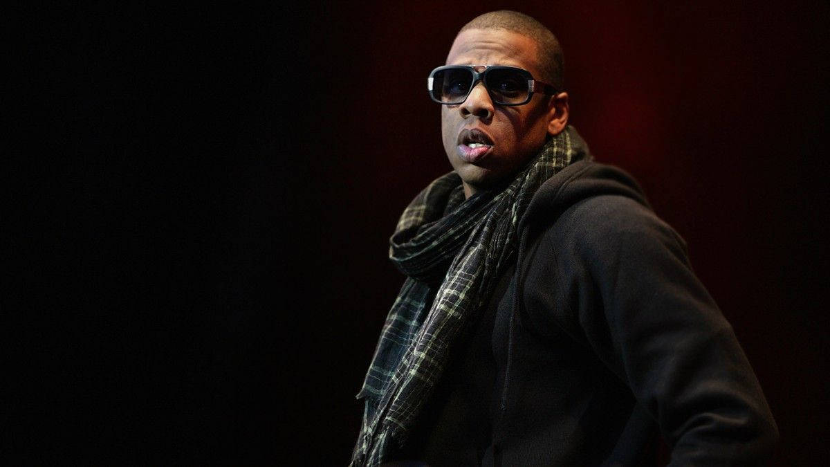 Jay-Z 美國饒舌歌手 壁紙 — 一名戴著太陽眼鏡和格子圍巾的男子站在深紅色背景前，燈光效果戲劇化。