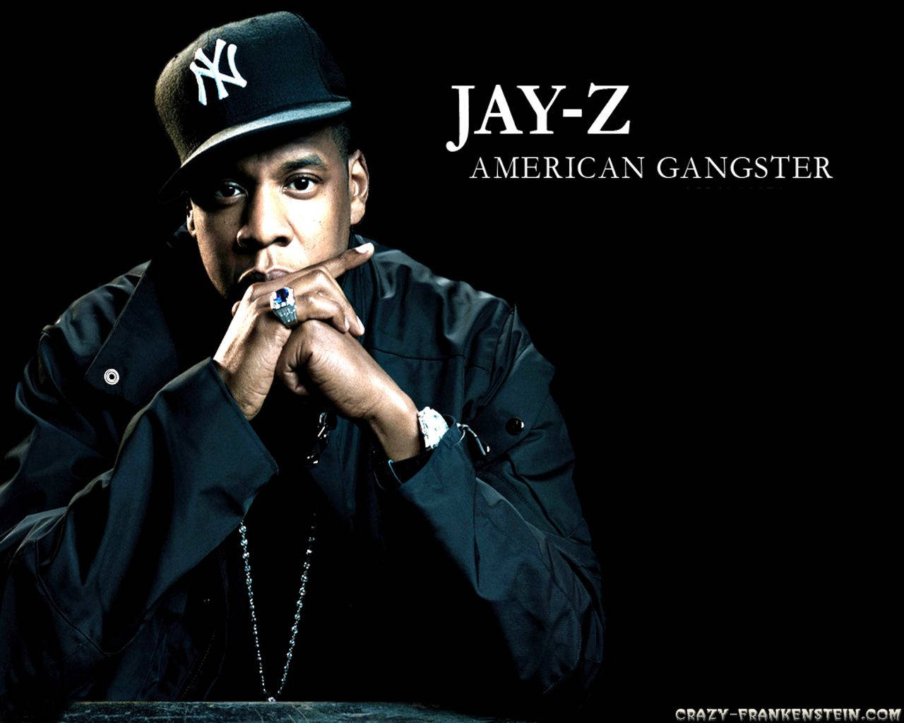 Jay-Z 美國黑幫 壁紙 — Jay-Z 凝視著，雙手緊握在嘴邊，戴著NY帽子和黑色外套，背景為黑色。