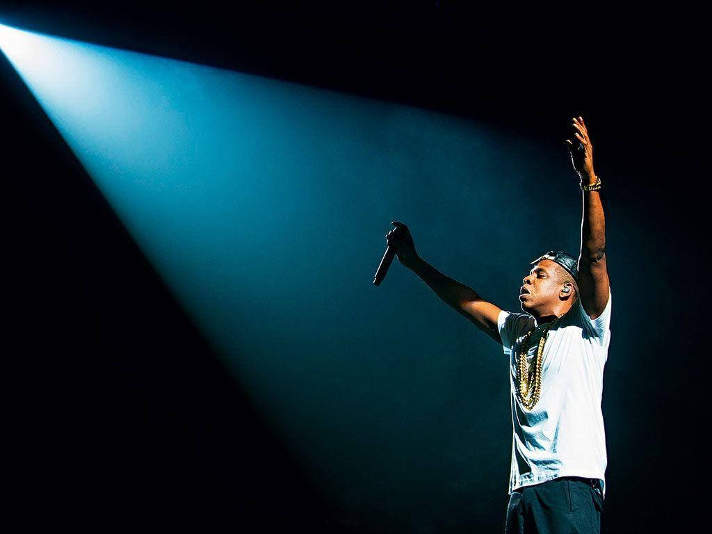 Jay-Z 被強光照射 壁紙 — 一位表演者沐浴在單一藍色聚光燈下，在黑暗的舞台上舉起手臂和麥克風。