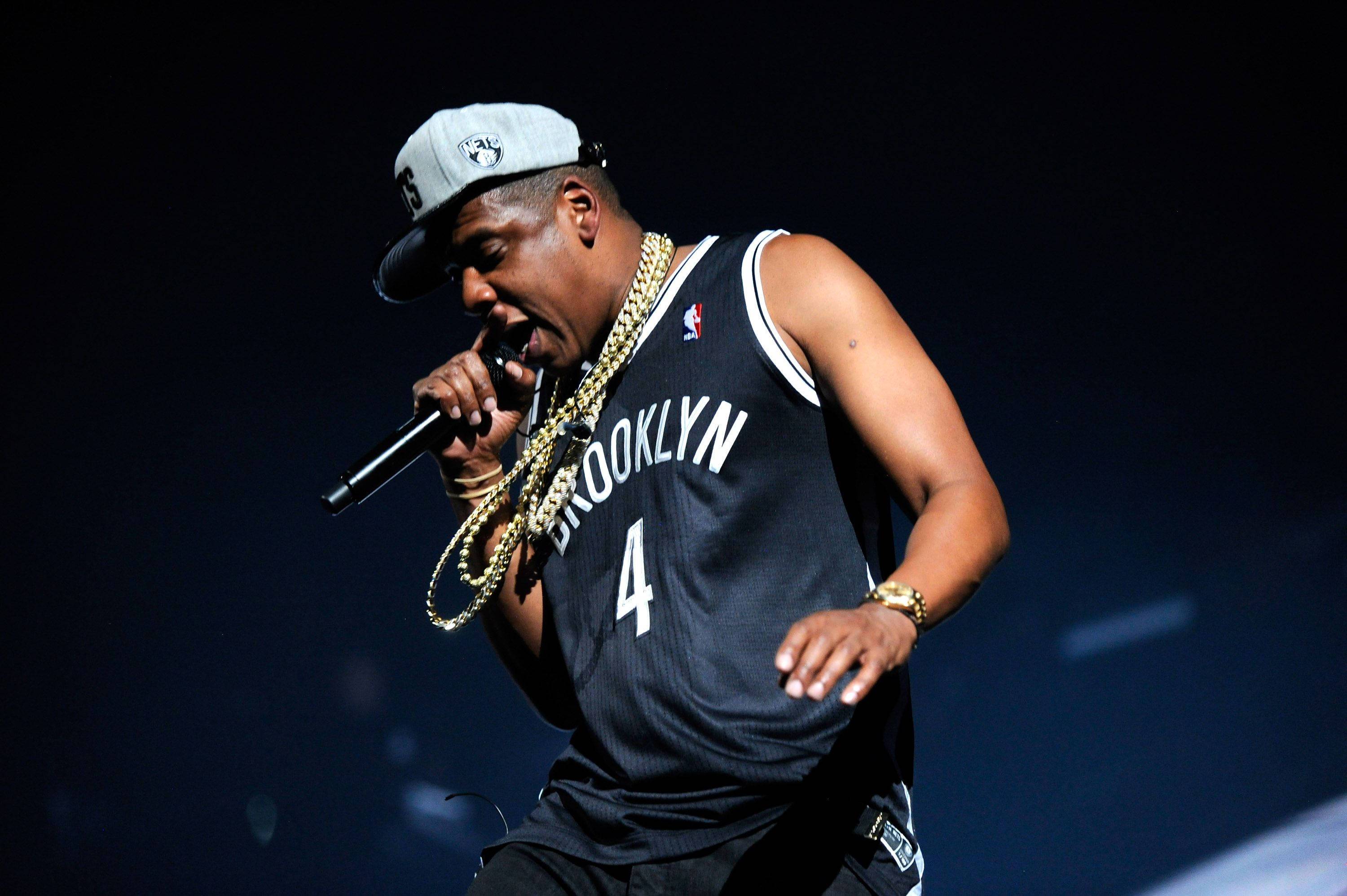 Jay-Z 身穿NBA球衣 壁紙 — Jay Z 在舞台上熱情演出，身穿布魯克林球衣和金鏈，在戲劇性的藍色燈光下。