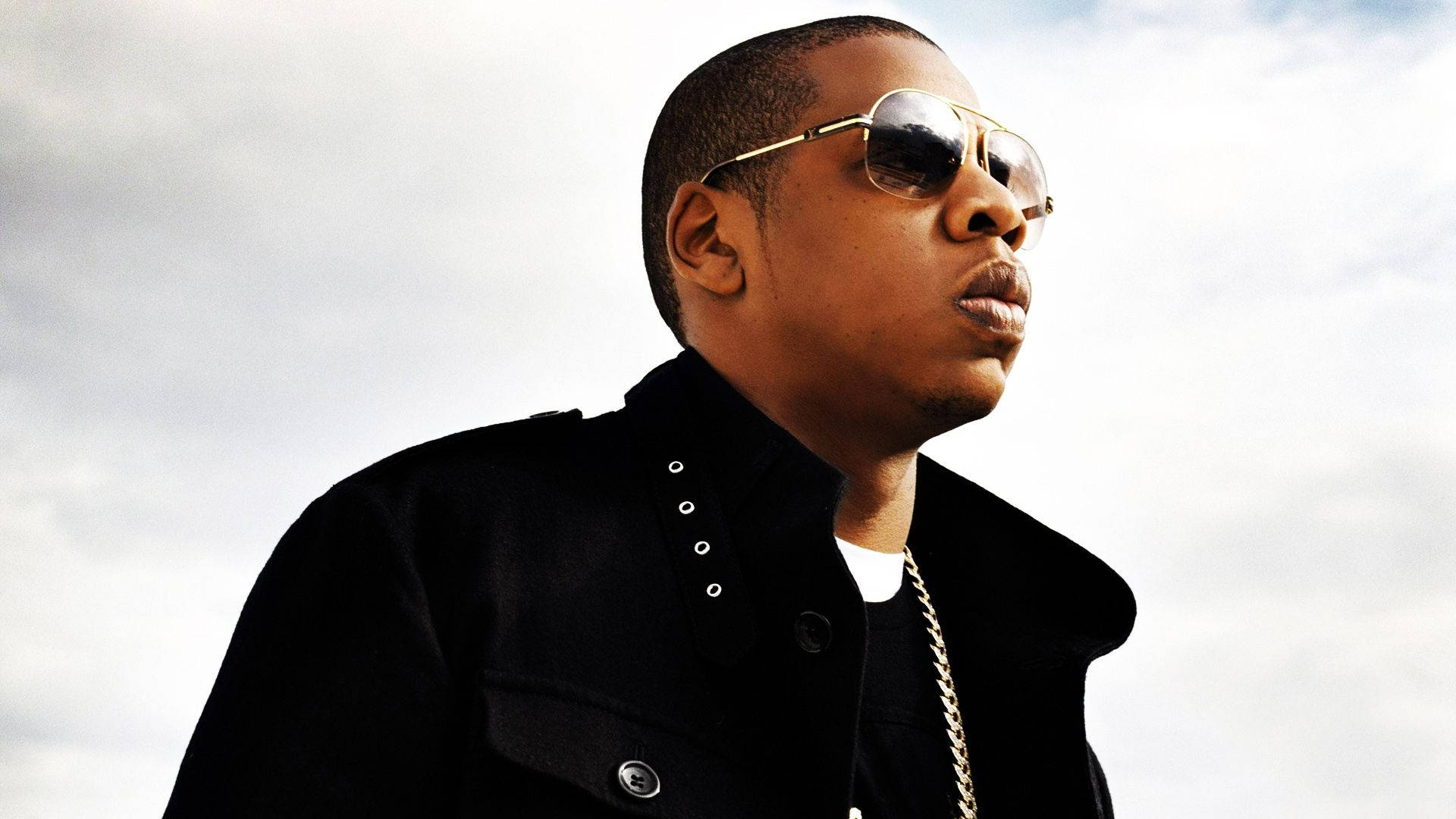 Jay-Z 雲天背景 壁紙 — 一名戴著飛行員太陽眼鏡和黑色外套的男子，在陰天自信地擺姿勢。
