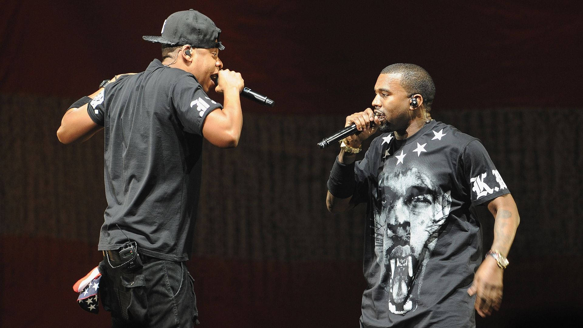 Jay-Z與Kanye West合唱 壁紙 — 兩位饒舌歌手在明亮的舞台燈光下手持麥克風充滿活力地表演。