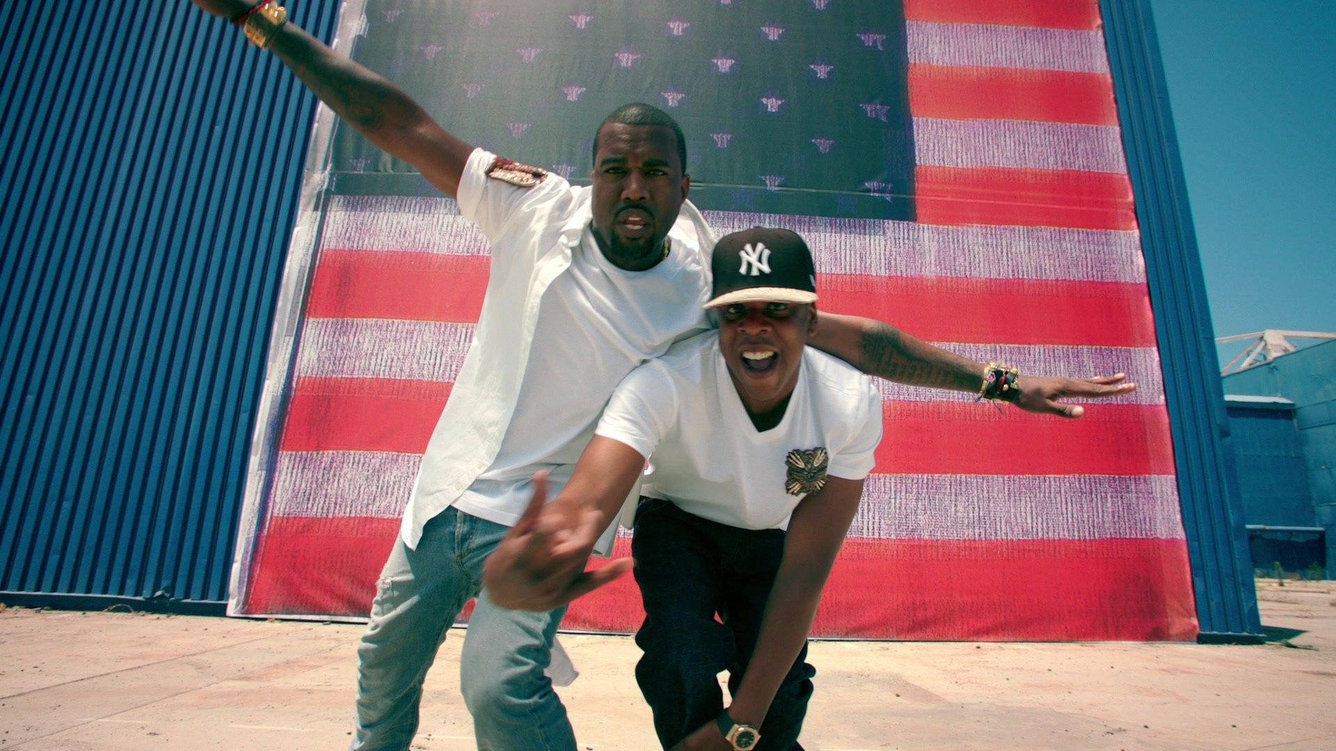 Jay-Z與Kanye West與美國國旗 壁紙 — 兩名男子在明亮的日光下，於藍色工業牆上巨大的美國國旗壁畫前動態擺姿勢。