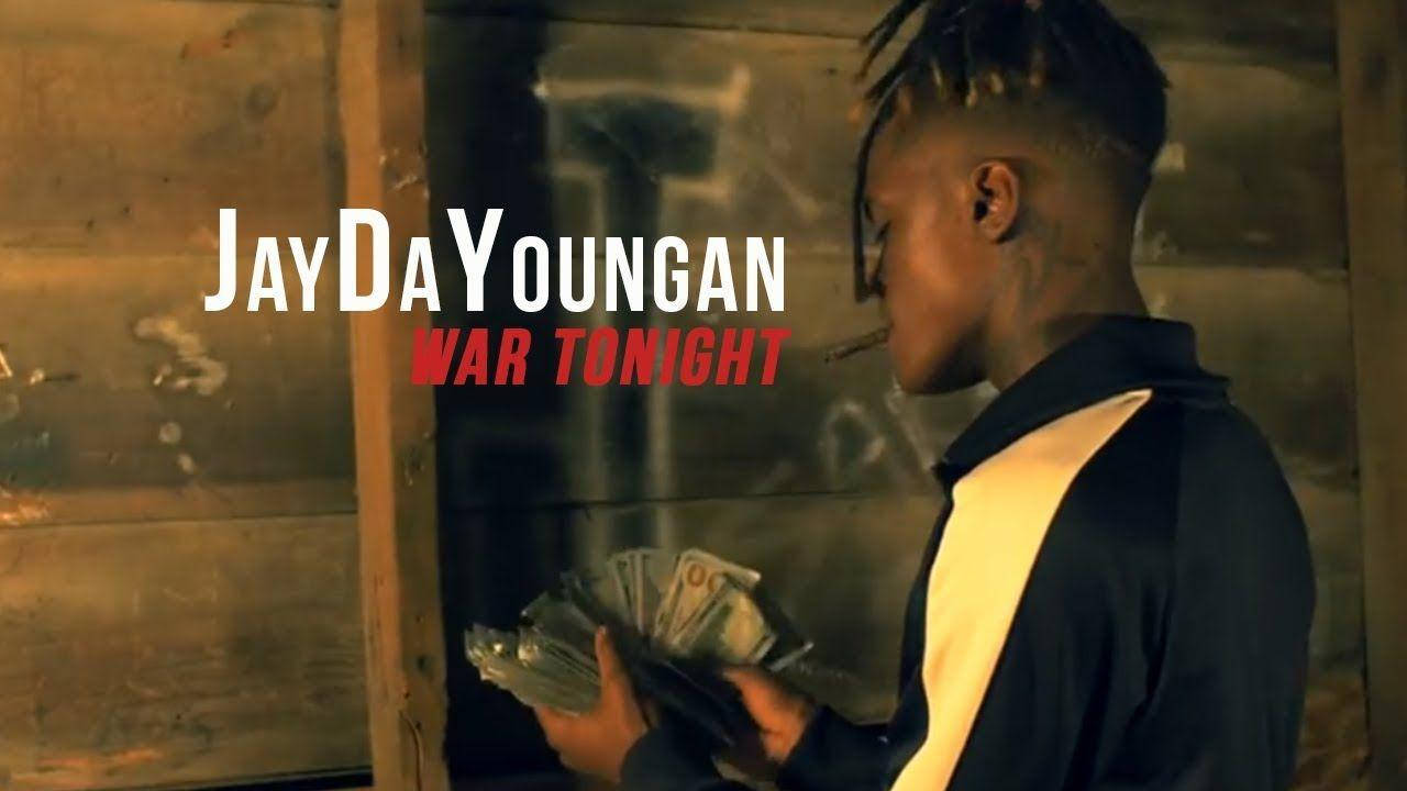 JayDaYoungan《War Tonight》歌曲照 壁紙 — 一個年輕的雷鬼辮男子在溫暖燈光下，靠著風化的木牆抽煙，同時手持現金。