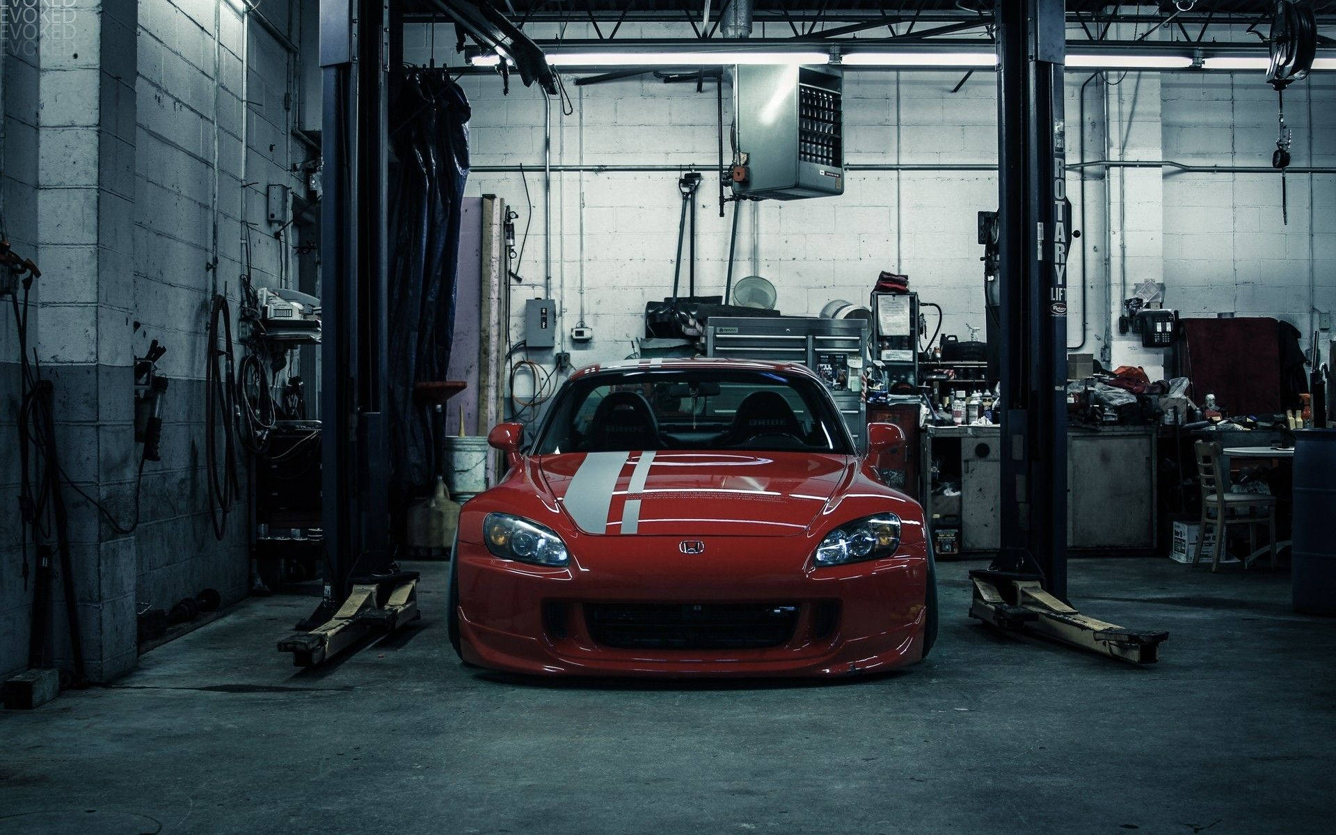 紅色JDM本田S2000車庫 壁紙 — 一輛帶有白色賽車條紋的紅色跑車位於昏暗、雜亂的汽車修理廠的升降機中央。