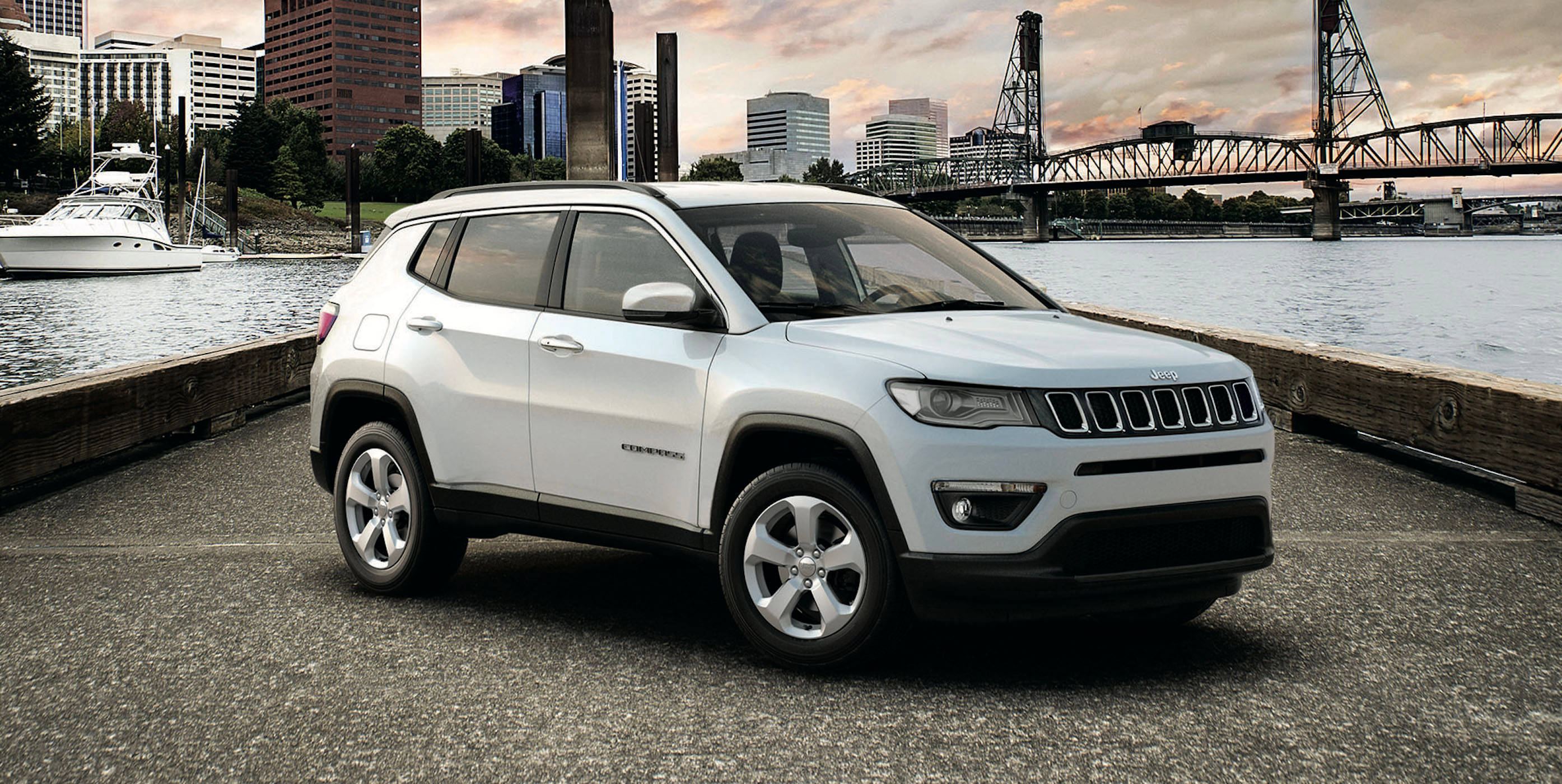 奢華優雅 — 白色 Jeep Compass 壁紙 — 一輛白色Jeep Compass停泊在海濱碼頭，背景是日落時分的城市天際線和橋樑。