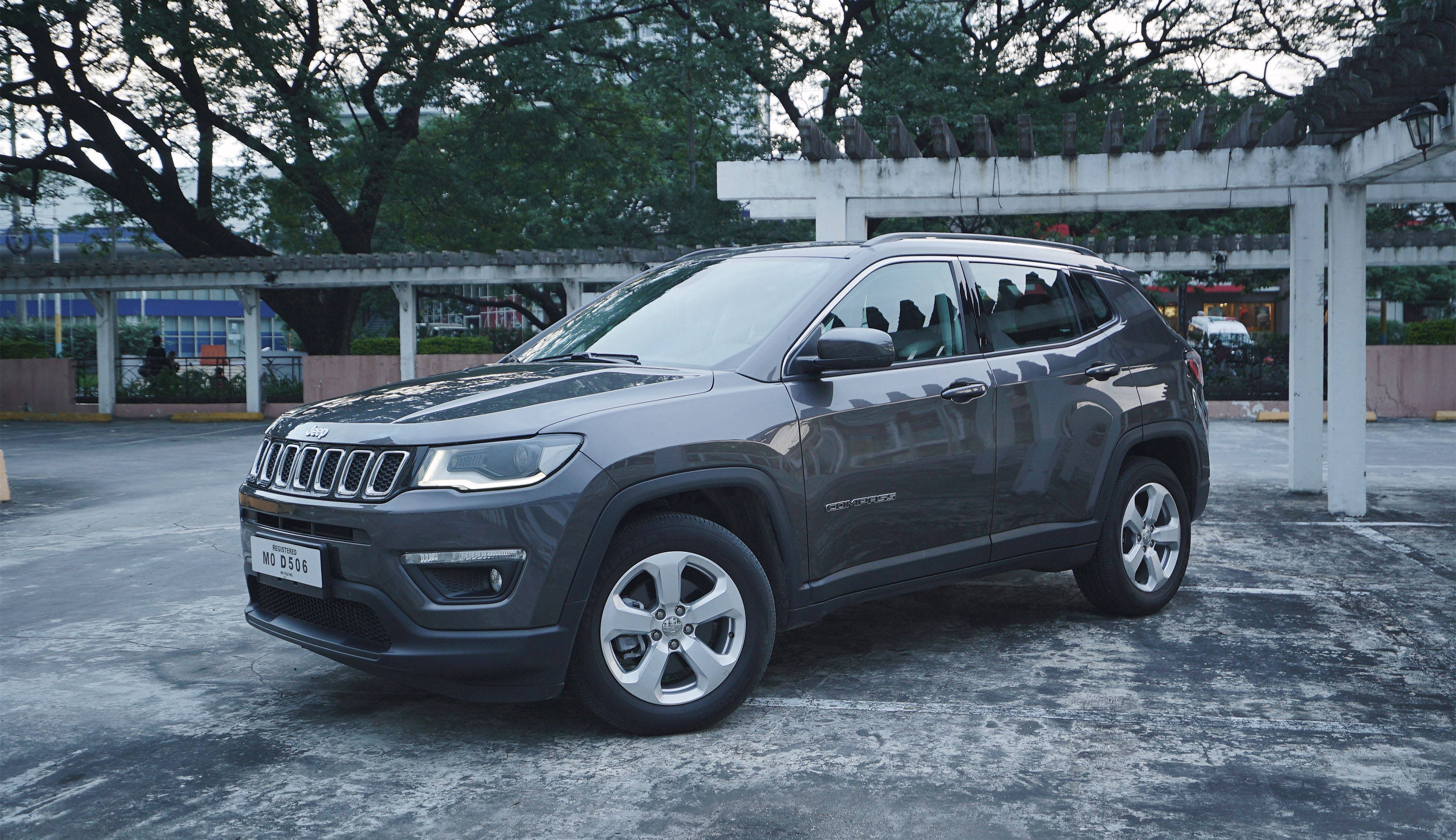 Jeep Compass停泊於灰色水泥地面 壁紙 — 一輛時尚的灰色Jeep Compass SUV停泊在樹蔭下的鋪砌停車場，環境光線柔和。