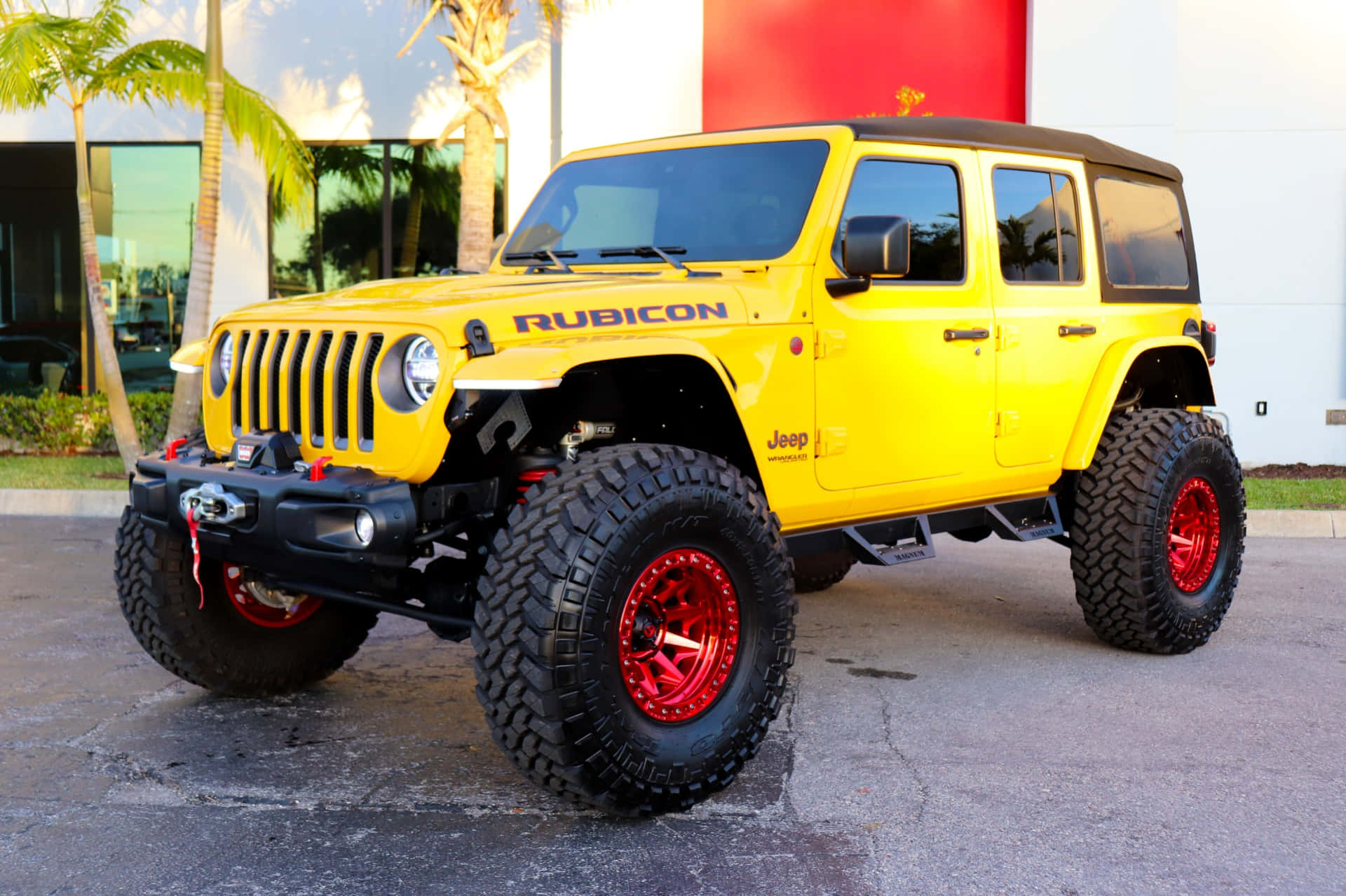 Jeep Wrangler：無所不至，無所不能 壁紙 — 一輛亮黃色Jeep Rubicon配備紅色輪圈和超大輪胎，停在棕櫚樹和現代建築附近的瀝青路面上。
