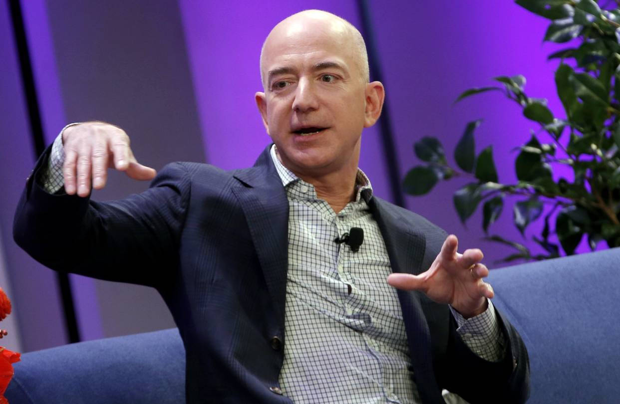 Bezos 桌布 壁紙 — Jeff Bezos 坐在藍色沙發上，在充滿綠色植物的鮮豔紫色背景下生動地做手勢。