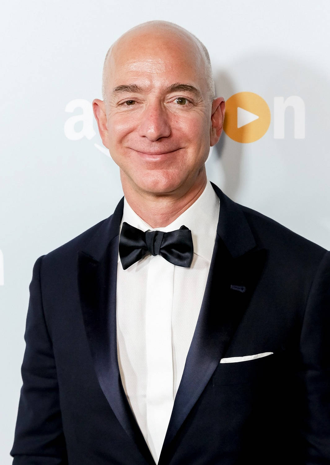 Bezos 桌布 壁紙 — Jeff Bezos 穿著俐落的黑色燕尾服配蝴蝶結，在淺灰色 Amazon Prime 背景下溫暖微笑。