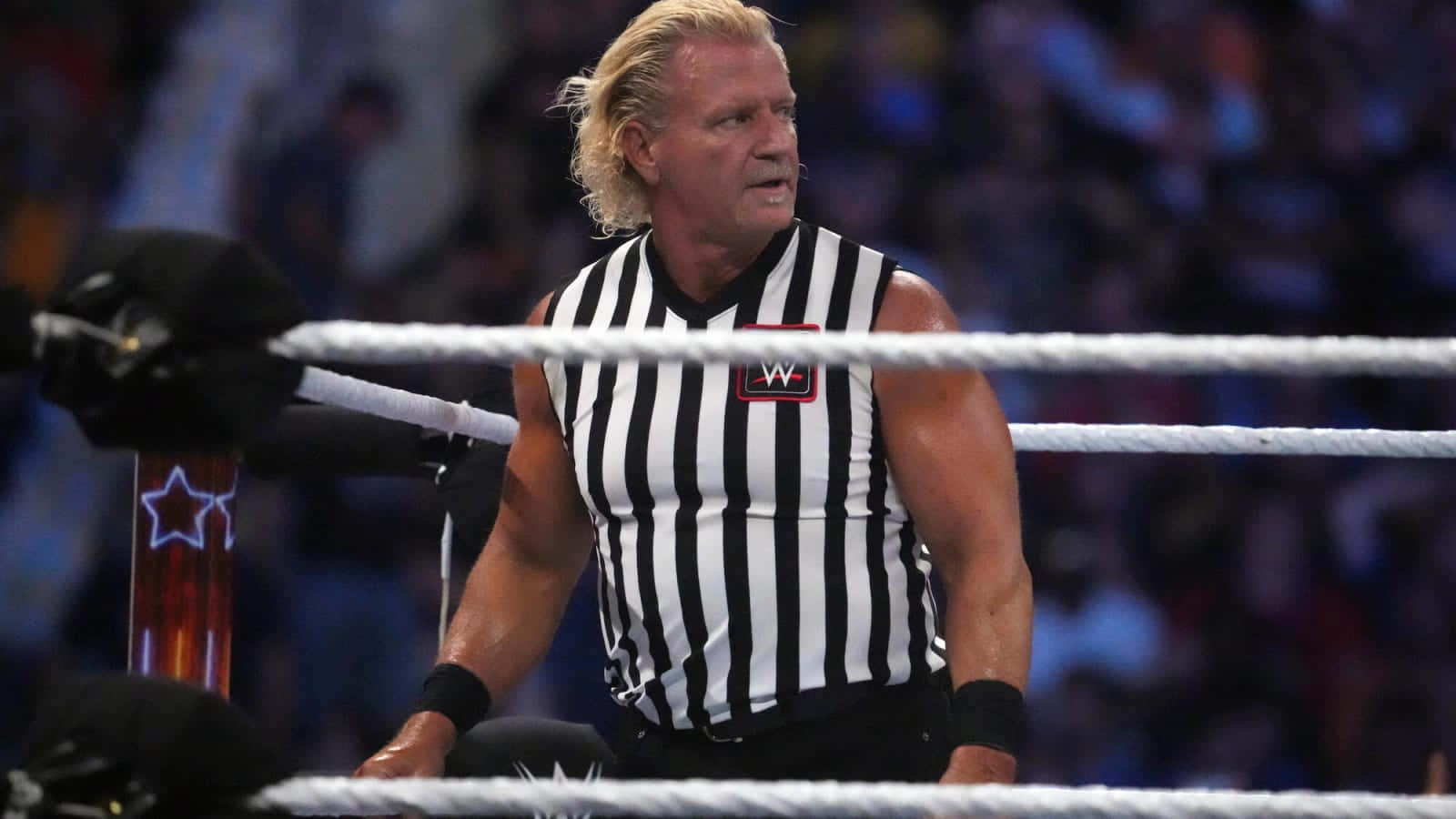 Jeff Jarrett WWE比賽攝影 壁紙 — 一位肌肉發達的WWE裁判穿著黑白間條衫，在明亮的場館燈光下，於摔角比賽中倚靠在擂台繩索上。