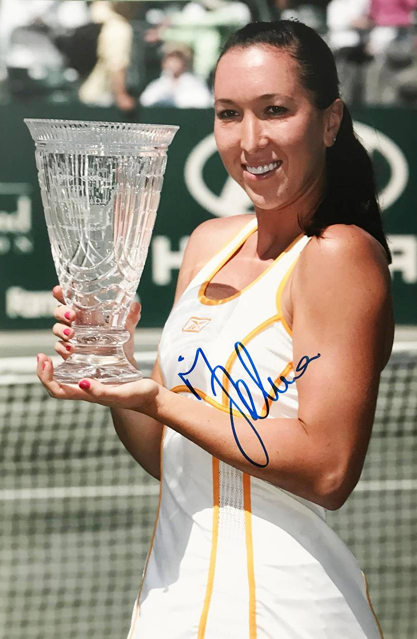 Jelena Janković手持查爾頓獎盃 壁紙 — 一位身穿白色和黃色連衣裙的網球選手微笑著，在陽光明媚的球場上舉起一個大型水晶獎盃。