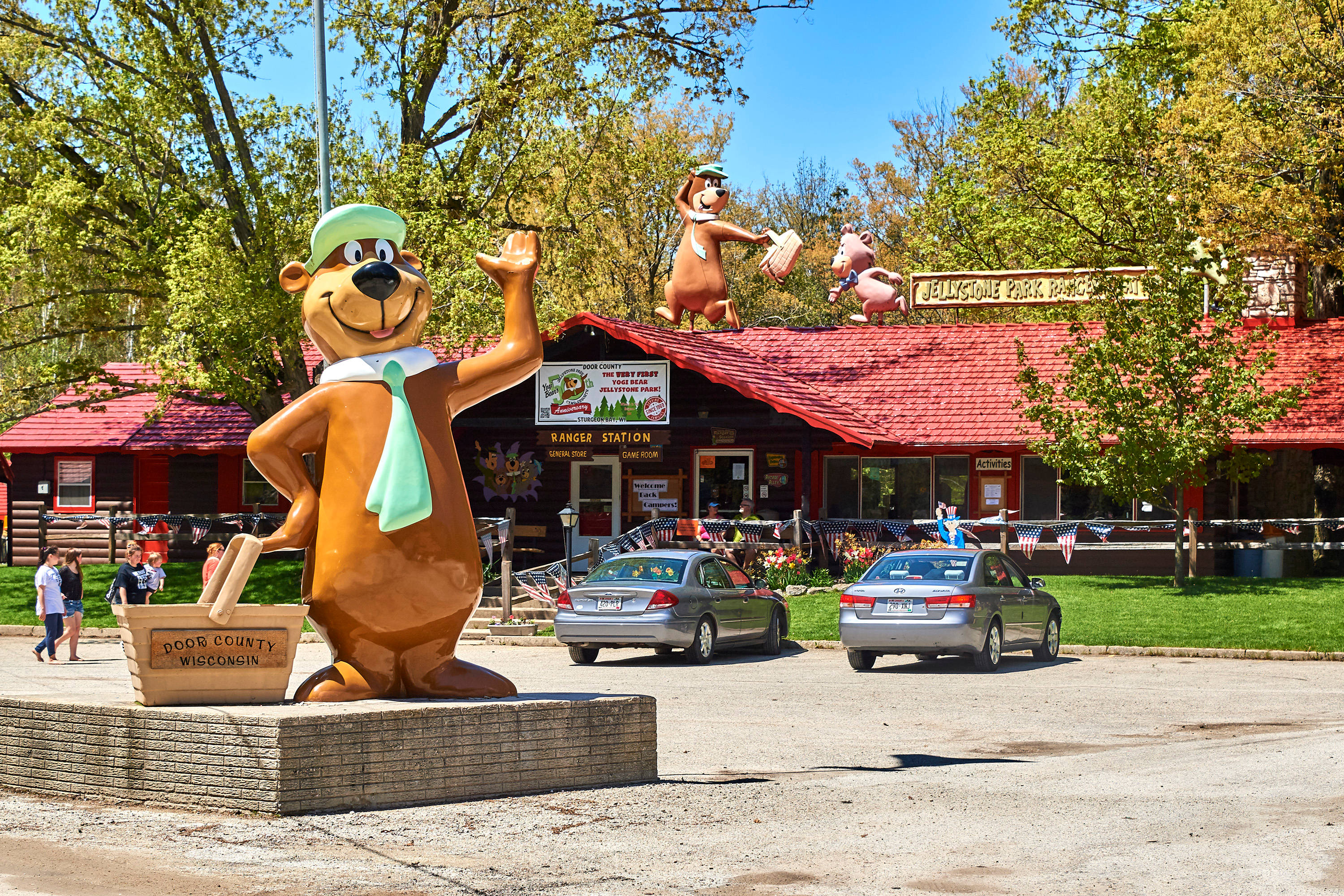 Jellystone 國家公園的 Yogi Bear 巨型雕像 壁紙 — 一個巨大的卡通海狸雕像在紅色屋頂的護林站旁揮手，前面有汽車停泊在晴朗的天空下。